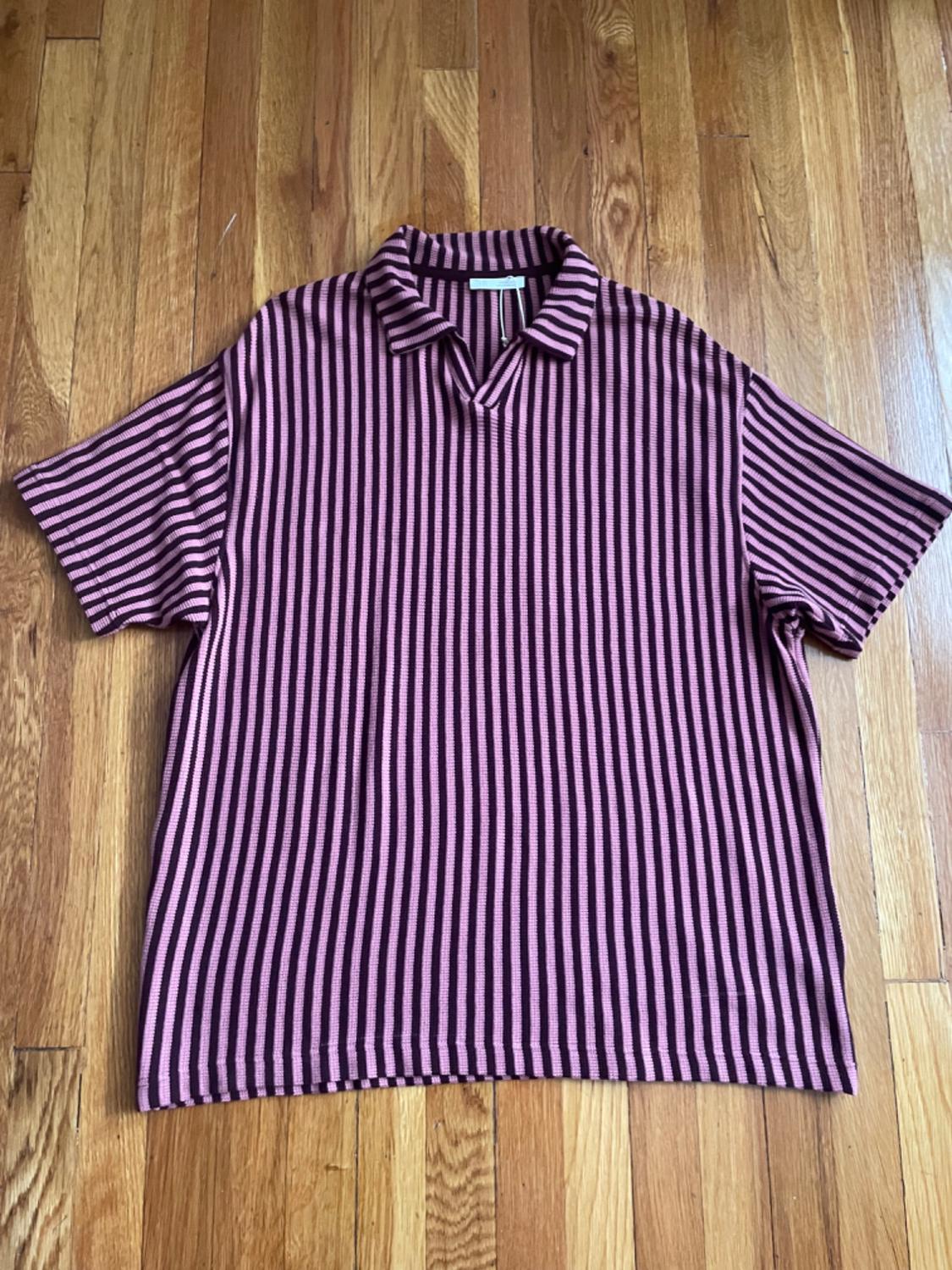 Short-sleeved polo shirt with dark and pink vertical stripes, with an open collar, laid out on a wooden floor.