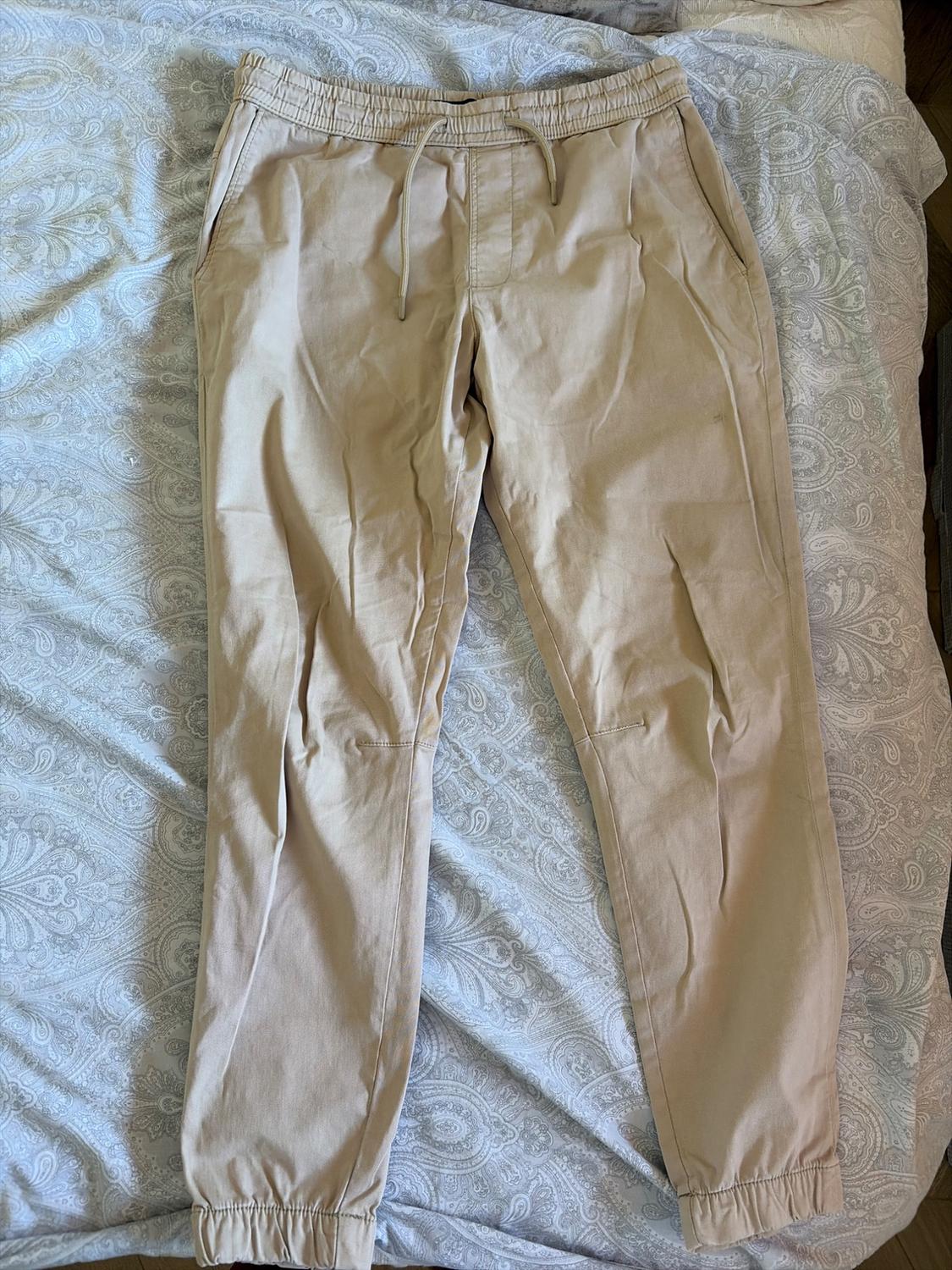 Beige trousers with an elasticated waist and drawstring, placed on a light-coloured paisley patterned bedspread.