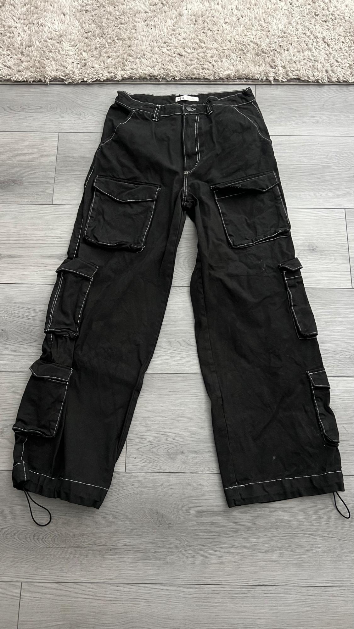 Black cargo pants with white stitching, multiple pockets, adjustable cuffs, and a ZARA tag, on a gray wooden floor.