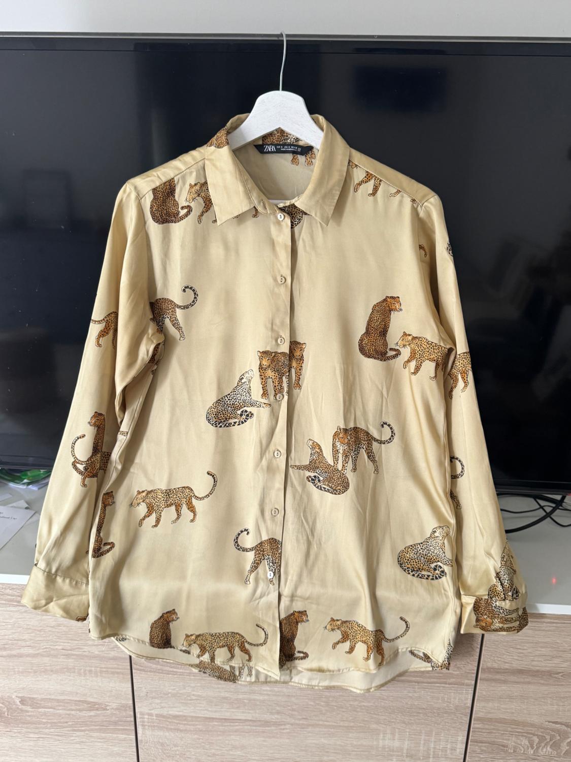 Beige shirt with leopard print in various positions, hanging on a white hanger in front of a black television.