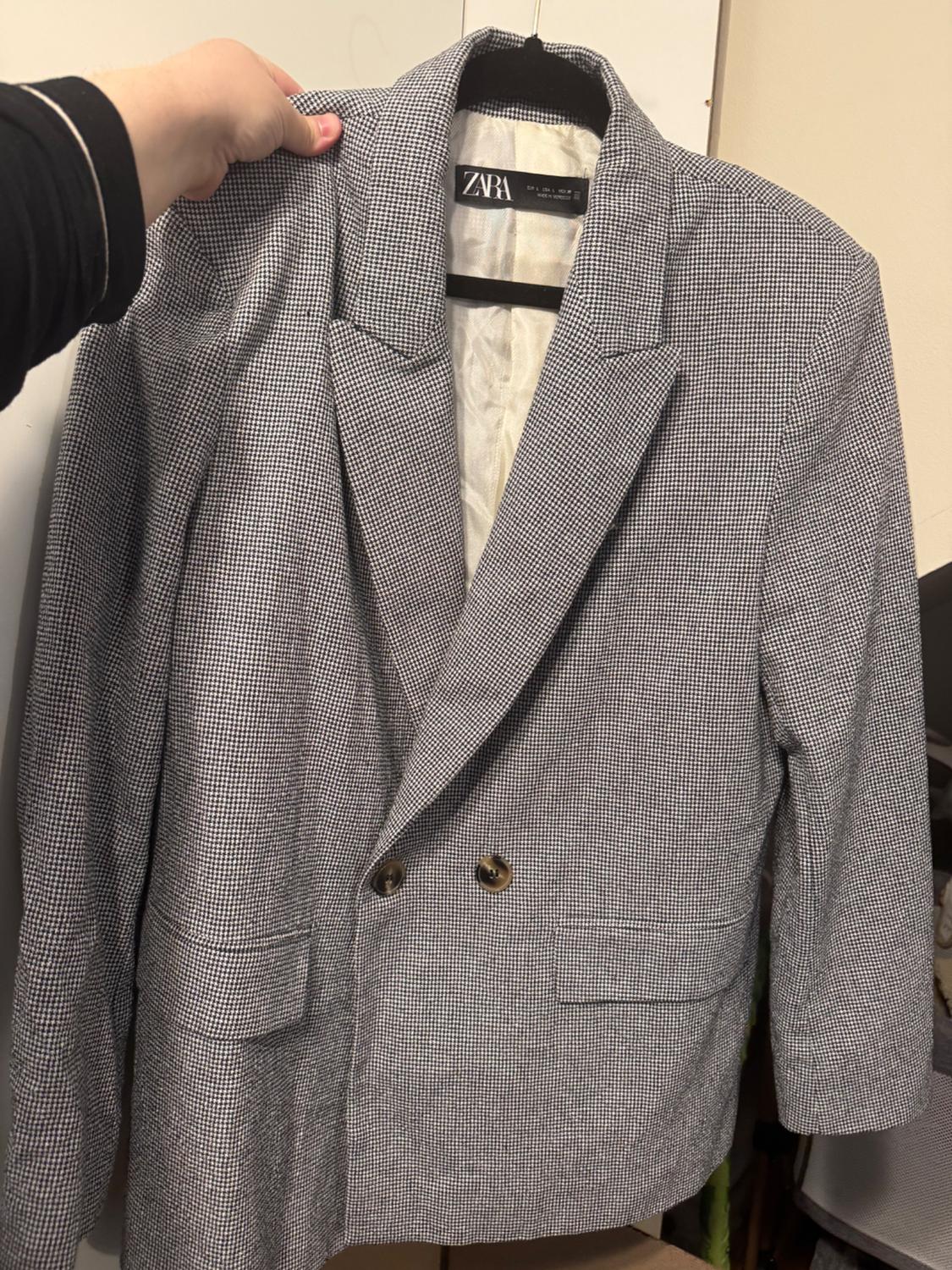 Hand holding a grey checked jacket with brown buttons, hanging on a hanger. Visible label: "ZARA".