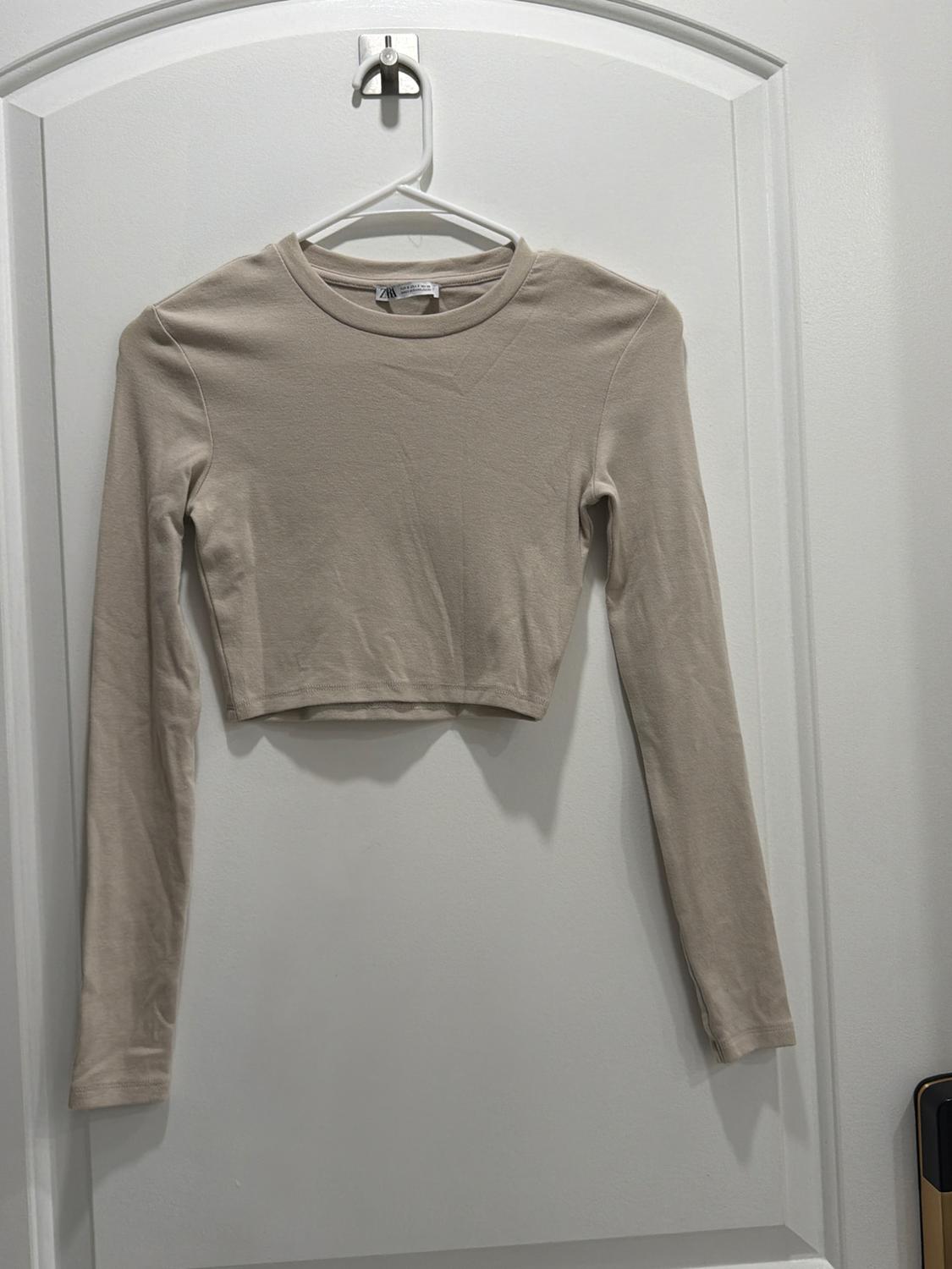 Beige long-sleeved cropped t-shirt hanging on a white hanger over a white door.