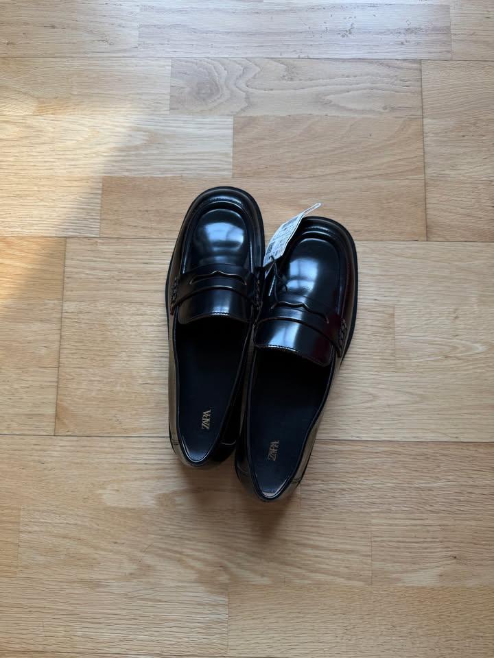 Shiny black loafers with a tag and the text ZARA on the insole, on a light wood floor.