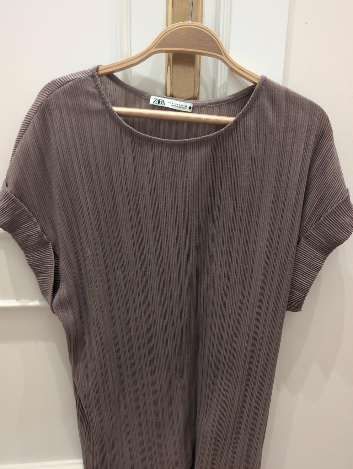 Brown pleated short-sleeved top on a wooden hanger over a white door. Text: ZARA EUR S USA S MEX 26.