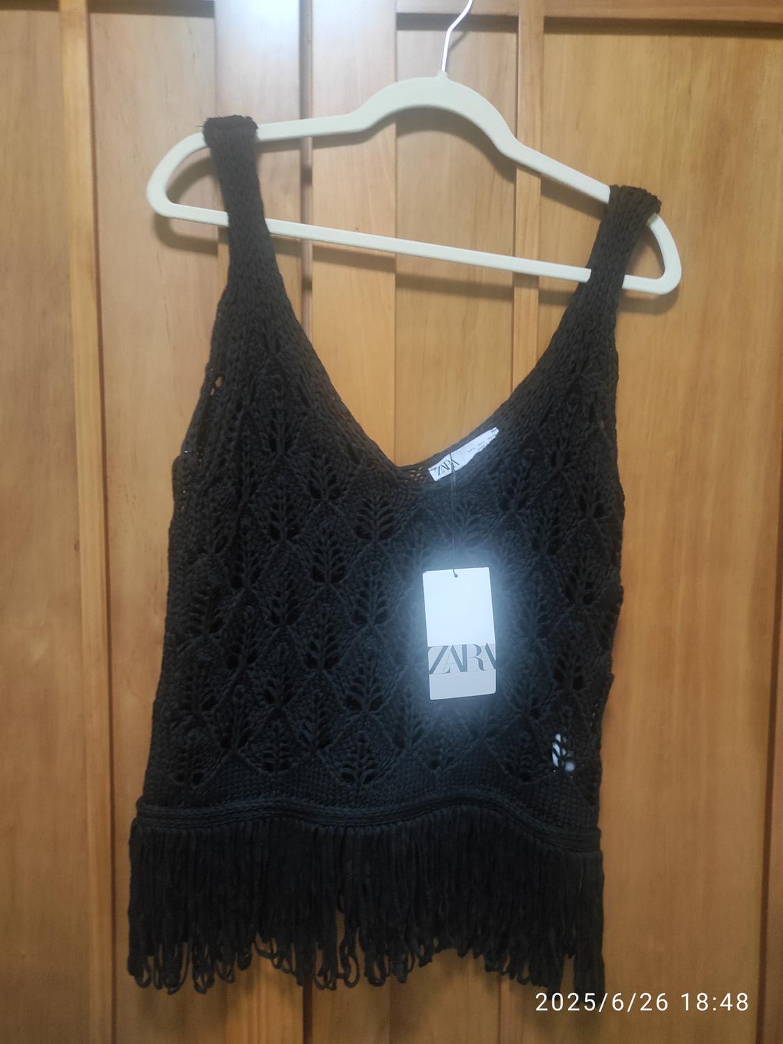 Black crochet top with fringing on the bottom edge, hanging on a white hanger in front of a wooden door. Zara logo visible on the label.