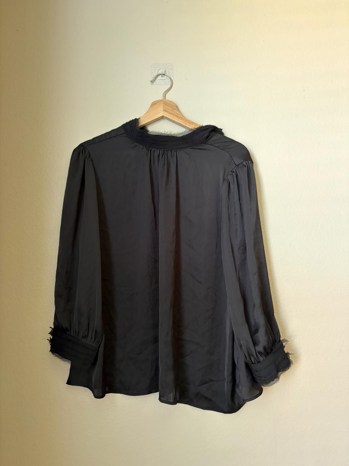 Lightweight black long-sleeved blouse with a ruffled collar and cuffs, hanging on a wooden hanger against a light-coloured wall. Simple and relaxed style.