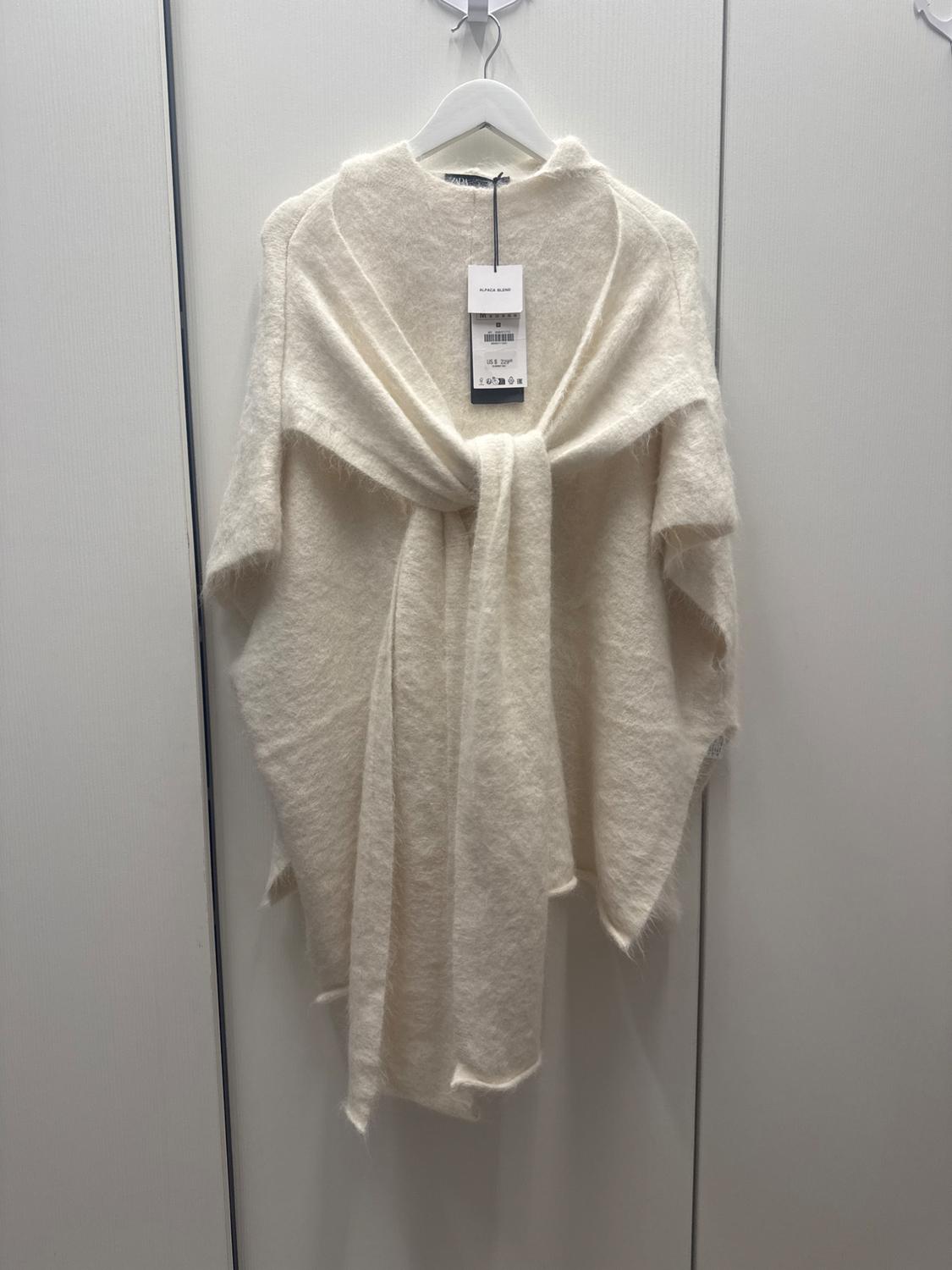 Beige wool poncho hanging on a white hanger, with a visible tag showing the price and product details. White background.