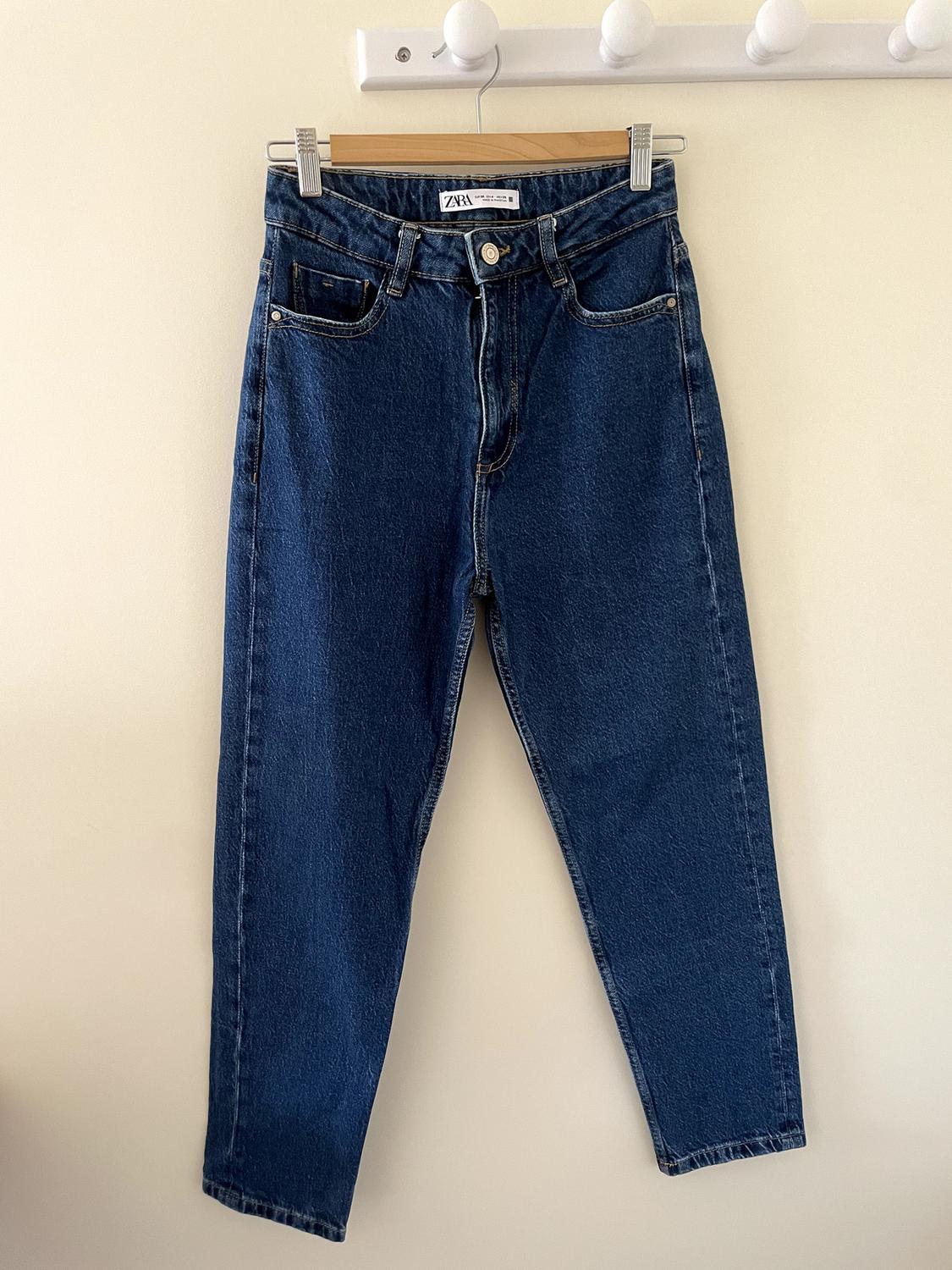Dark blue denim jeans hanging on a wooden hanger against a light wall. Label with the text ZARA.