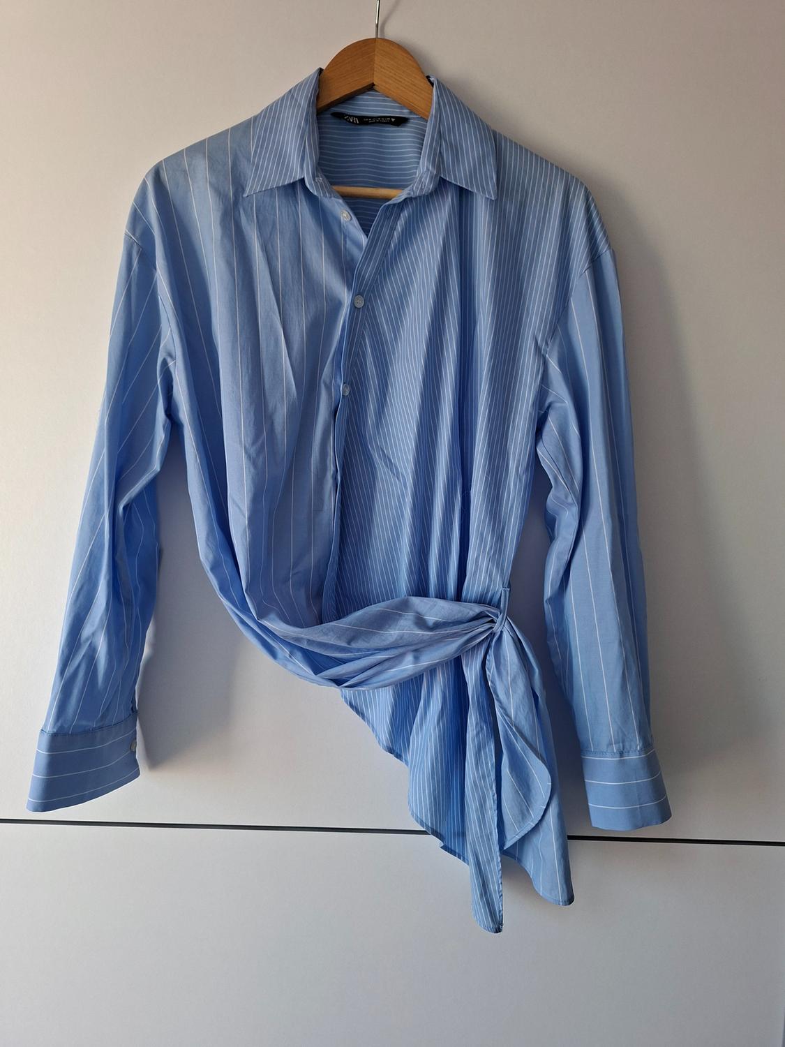 Long-sleeved blue shirt with white stripes hanging on a hanger against a white background. An asymmetrical knot at the bottom stands out.