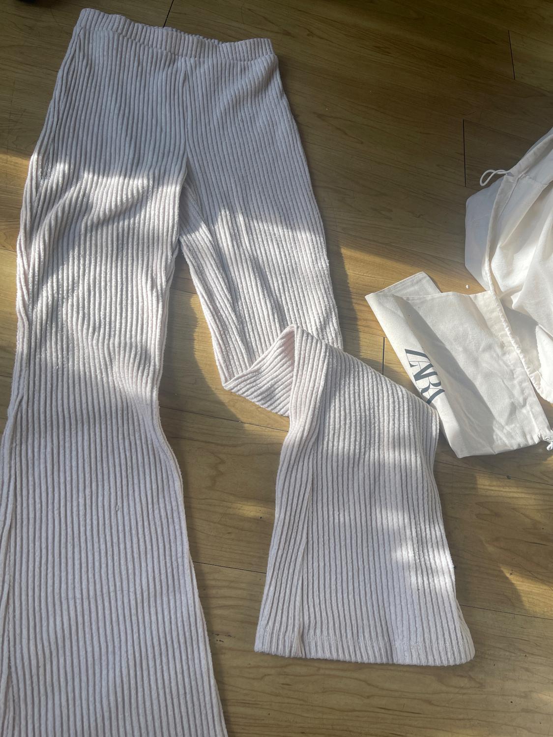 White ribbed pants on a wooden floor, with natural light. To the right, a white bag with partially visible text.