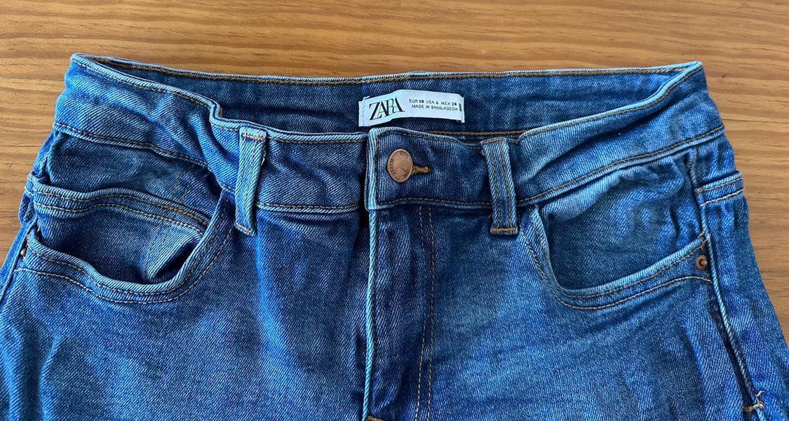 Close-up of the waistband of blue jeans on wood. Label with the text "ZARA EUR 38 USA 6 MEX 28 MADE IN BANGLADESH".
