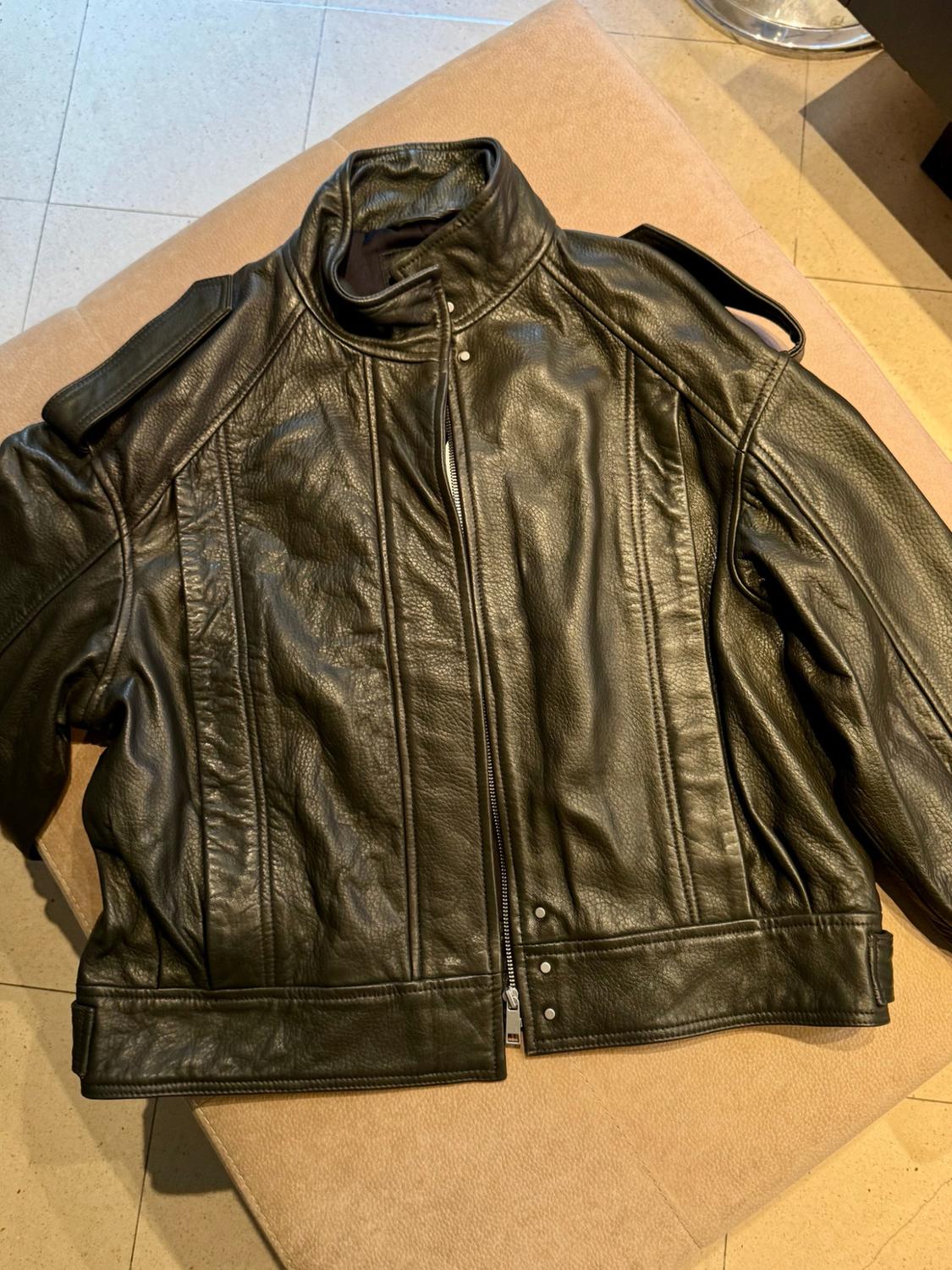 Black leather jacket with a front zip, stand-up collar and straps, resting on a beige seat with a tiled floor.
