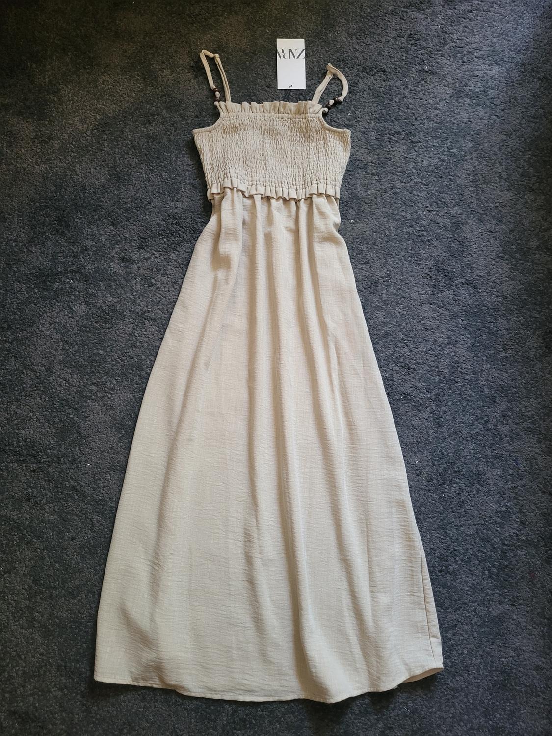 Beige maxi dress with spaghetti straps and a ruched bodice, on grey carpet. Label with text "AMZ" visible at the top.