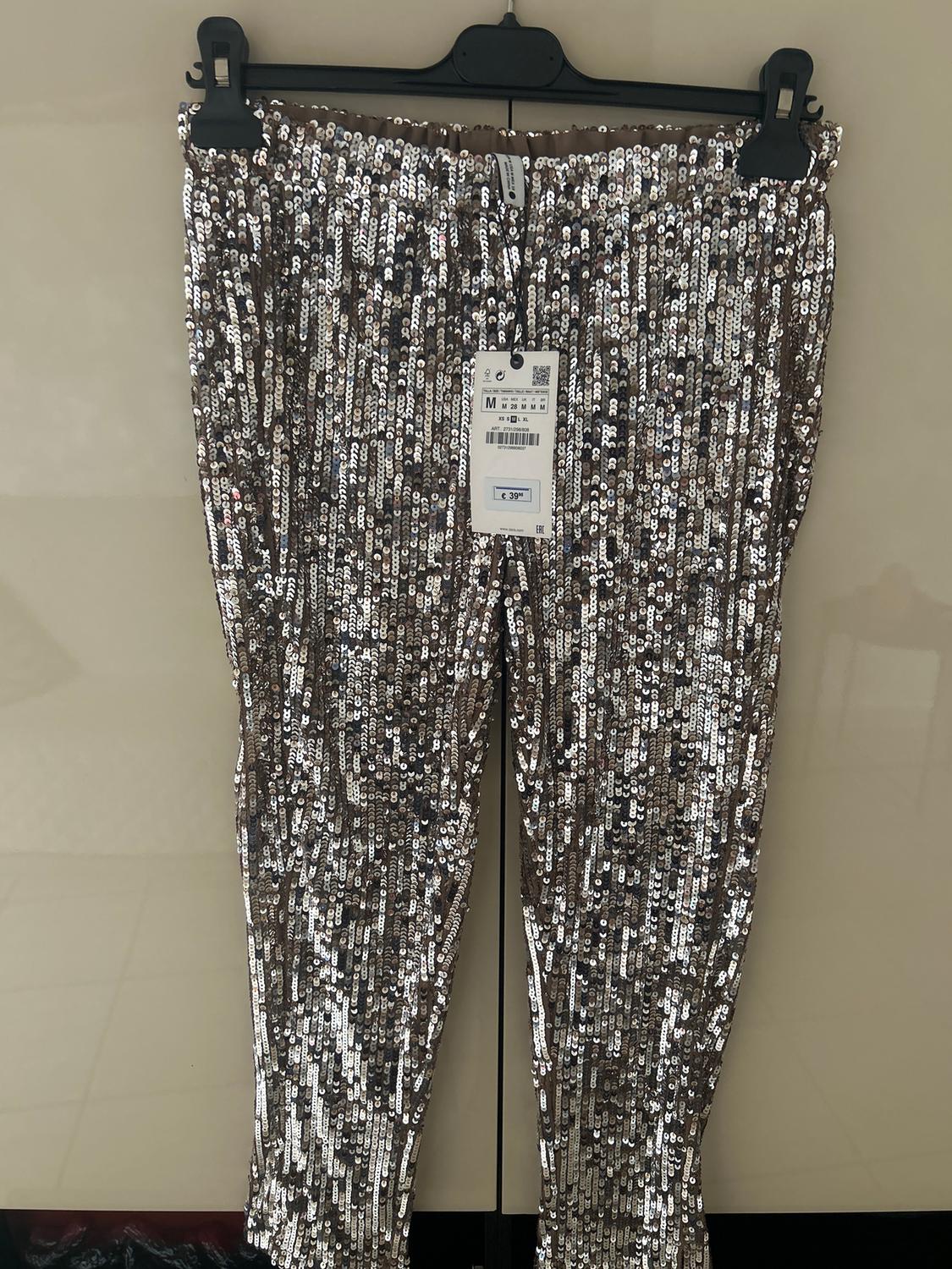 Silver sequinned trousers hanging on a black hanger. Visible tag with price of €39.99 and size M. Beige background.