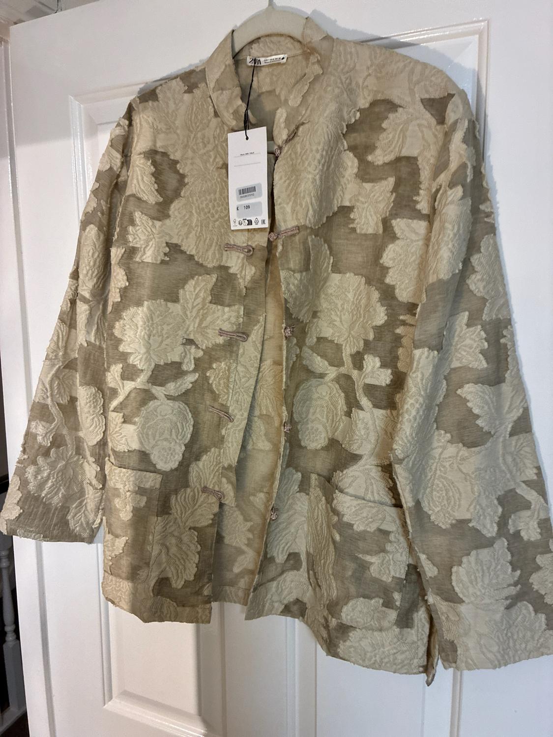 Beige jacket with embossed floral texture and knot fastenings, hanging on a white door. Label visible with the text £ 109.
