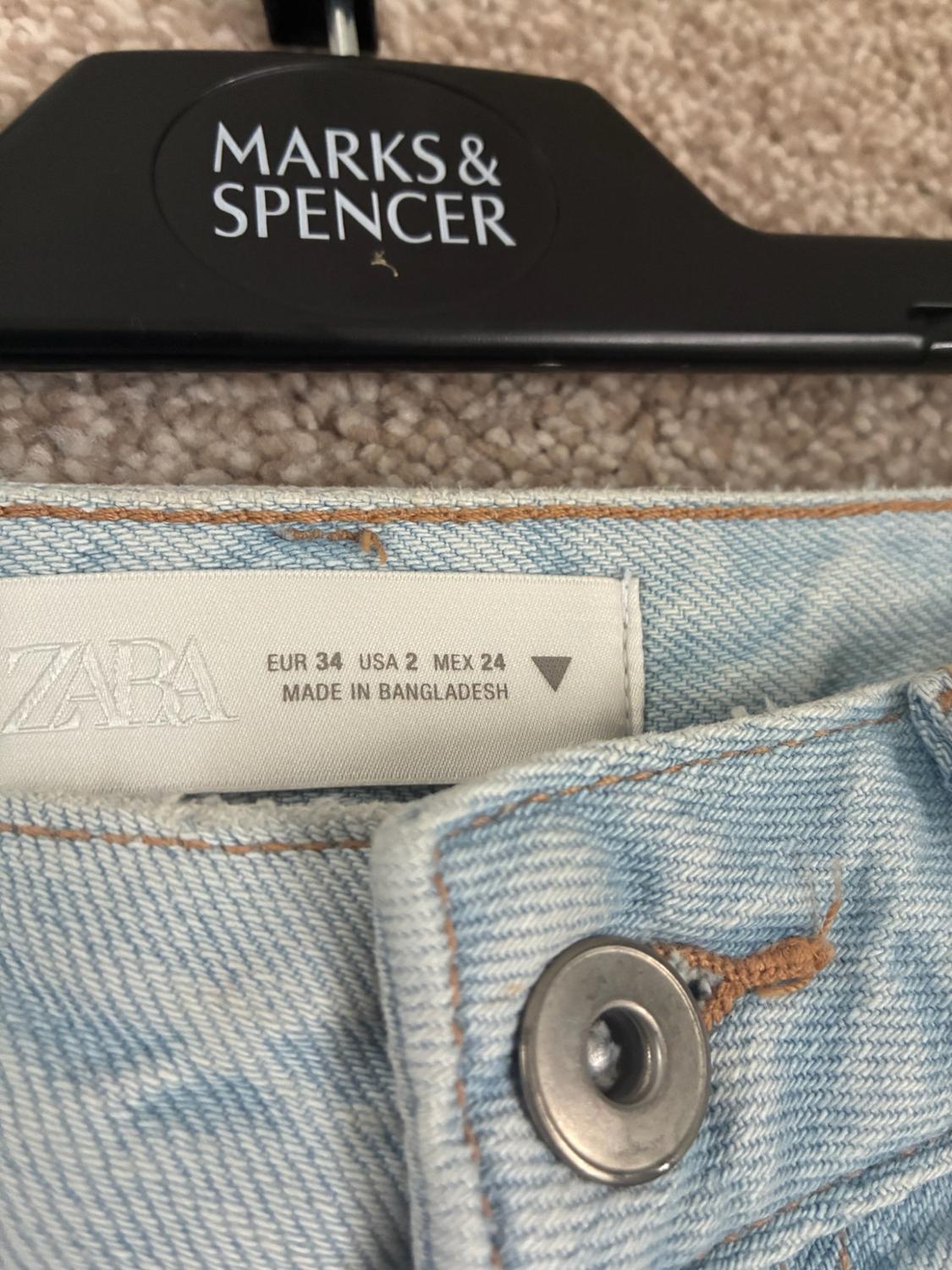 Close-up of jeans with a ZARA label reading EUR 34 USA 2 MEX 24 MADE IN BANGLADESH and a black hanger with the text MARKS & SPENCER.