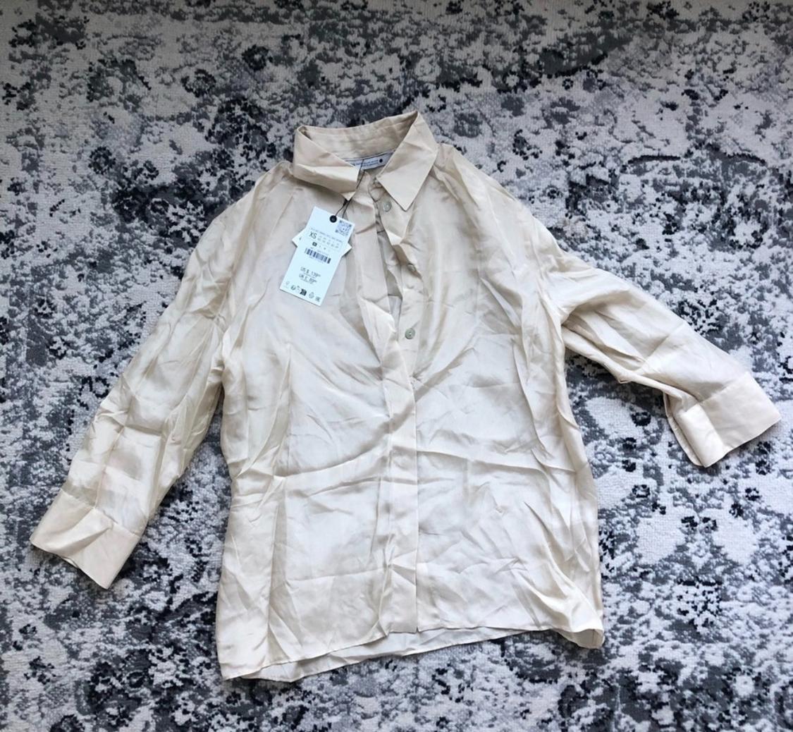Wrinkled long-sleeved beige shirt with a hanging tag, on an abstract-designed rug in gray and white tones.