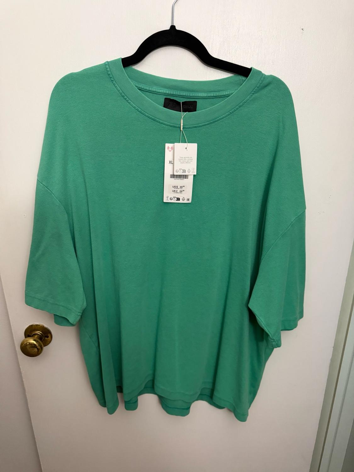Green short-sleeved t-shirt hanging on a white door. Label with text: XL, US $49.90, UK £29.99.