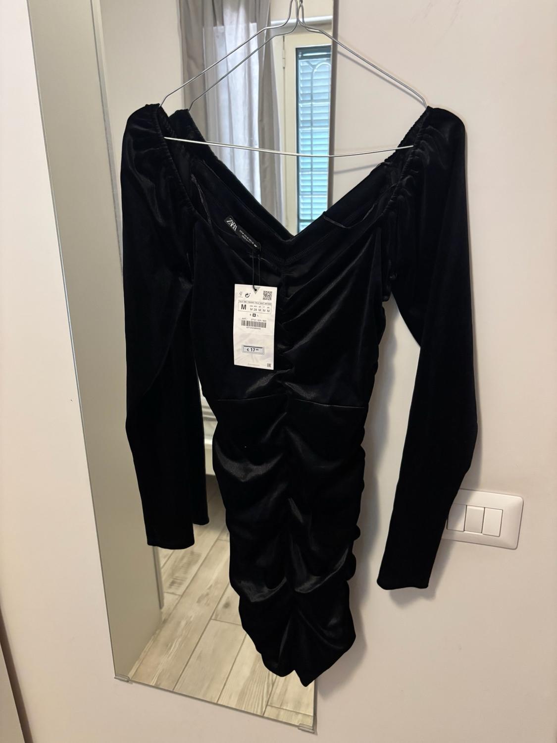 Short black gathered long-sleeved dress hanging on a hanger in front of a mirror. Includes tags bearing the text ZARA, M, €17.