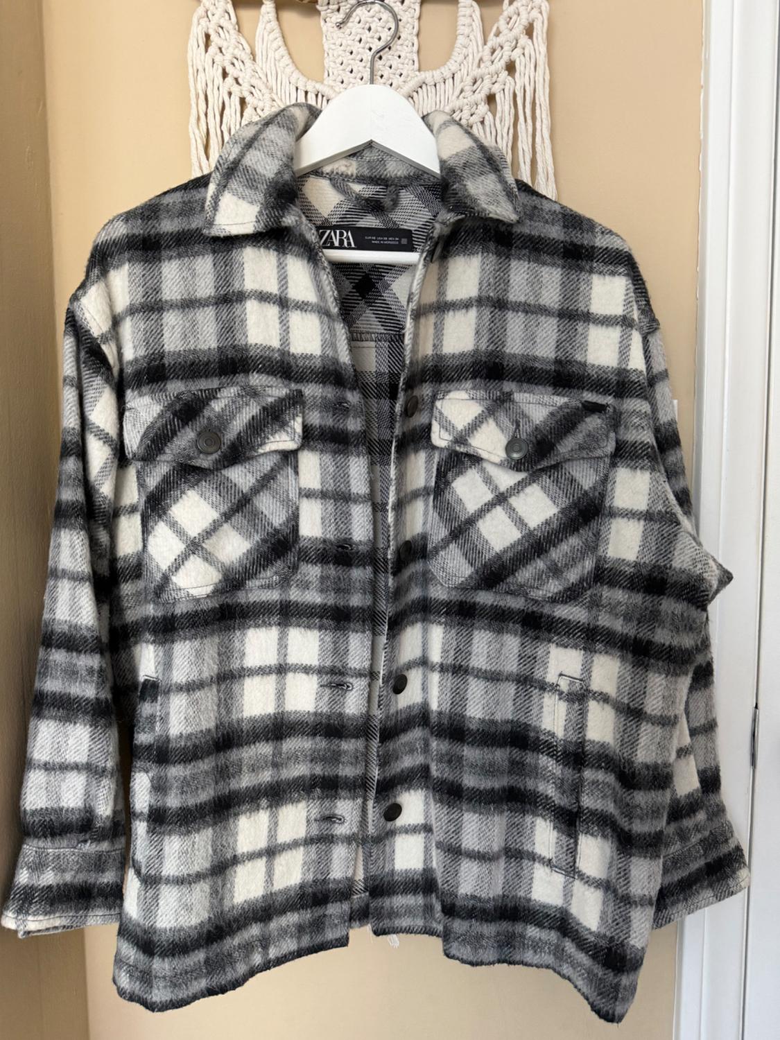 Black and white plaid shirt hanging on a white hanger. Beige background with macrame decoration. Visible tag: ZARA.