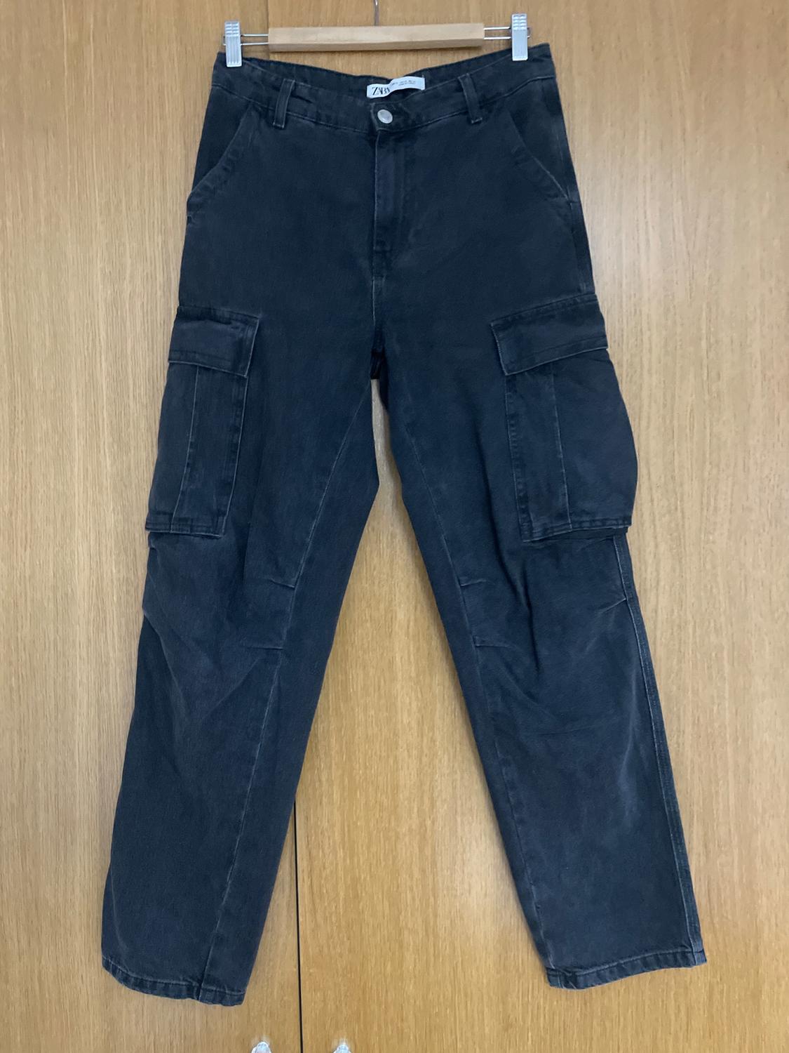Black cargo trousers hanging on a wooden hanger against a light wooden background. Large pockets on the sides.
