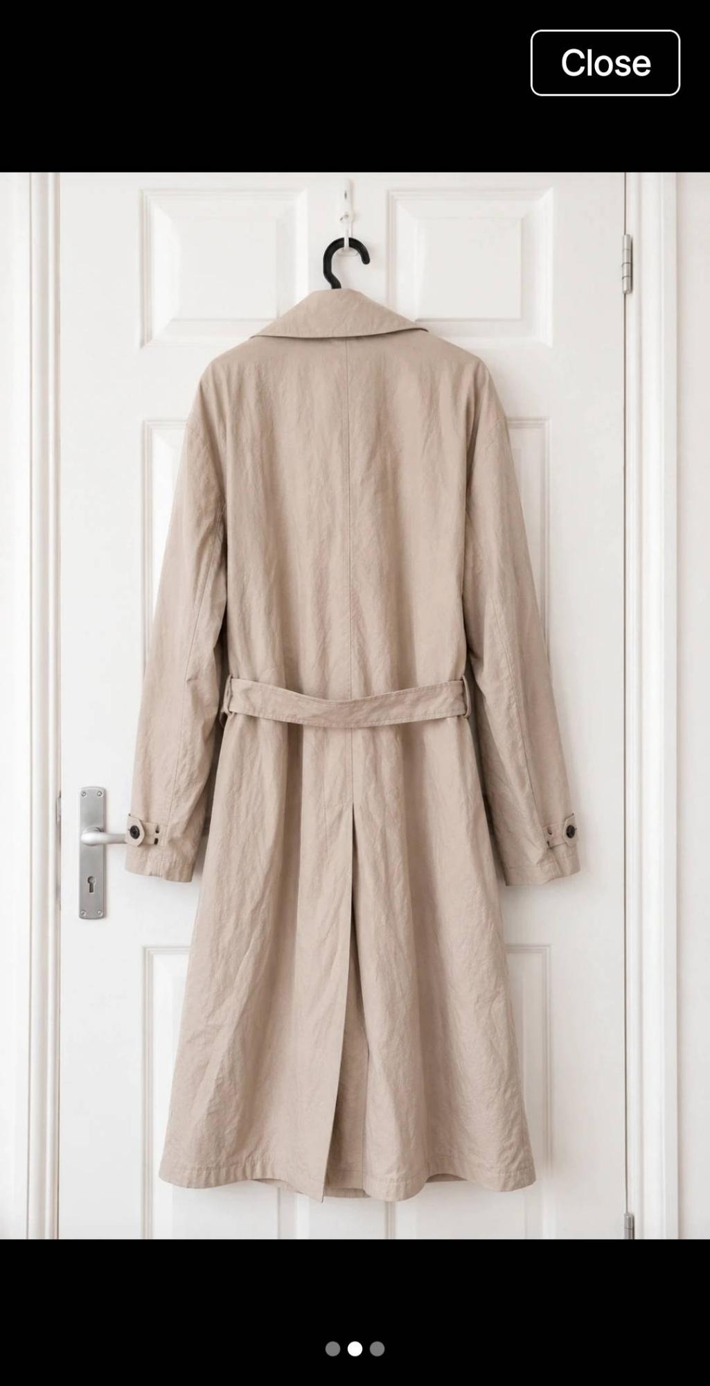 Beige trench coat seen from behind, hanging on a white door. It has a belt and a vent. In the top right, the text “Close” appears.