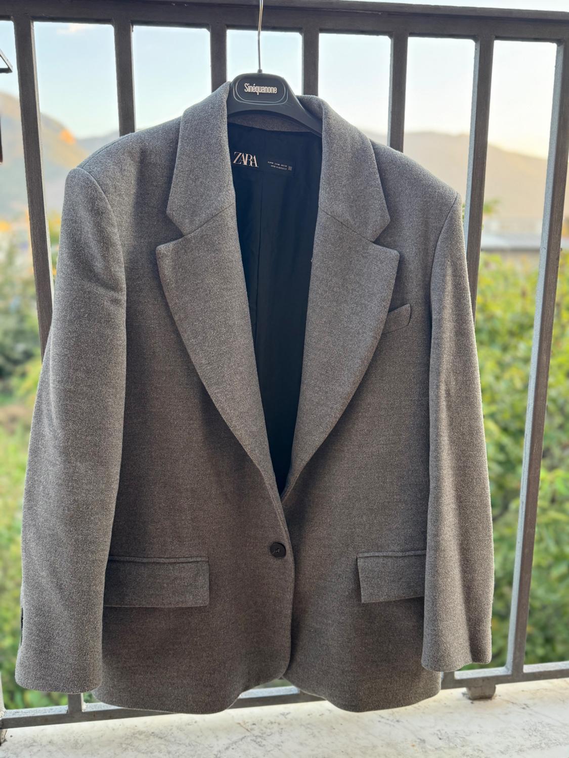 Grey jacket hanging on a black hanger with the label visible, on a balcony with a natural landscape background and clear sky.