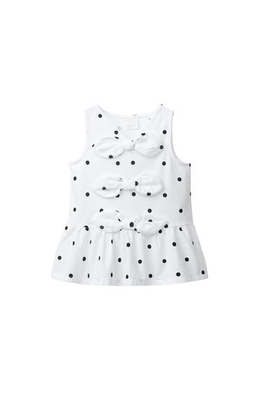Front view of a white sleeveless top with a black polka dot print, decorative ties on the front and a frilled hem.