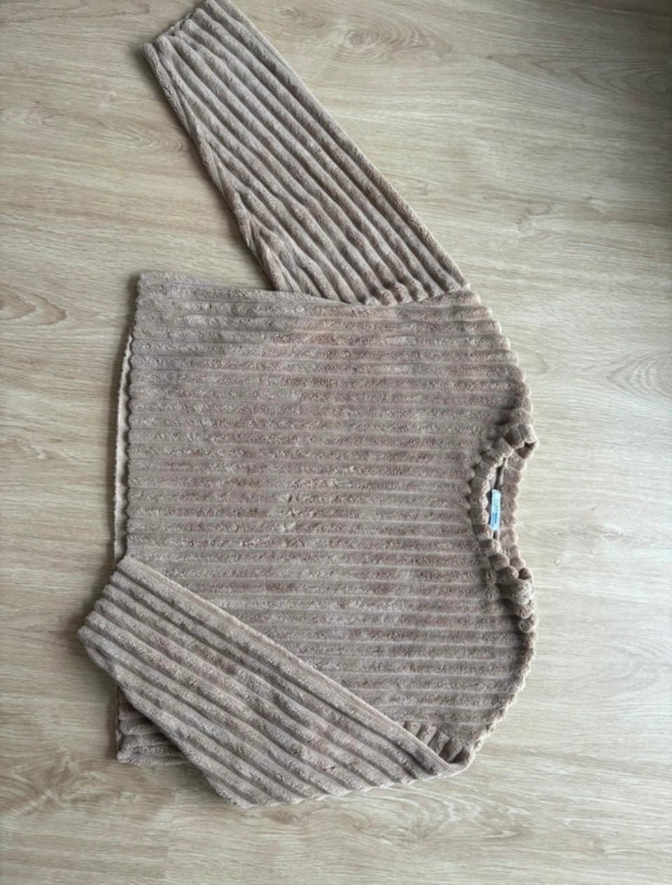 Beige long-sleeved jersey with a soft ribbed texture, folded on a light wooden floor. Relaxed and cosy style.