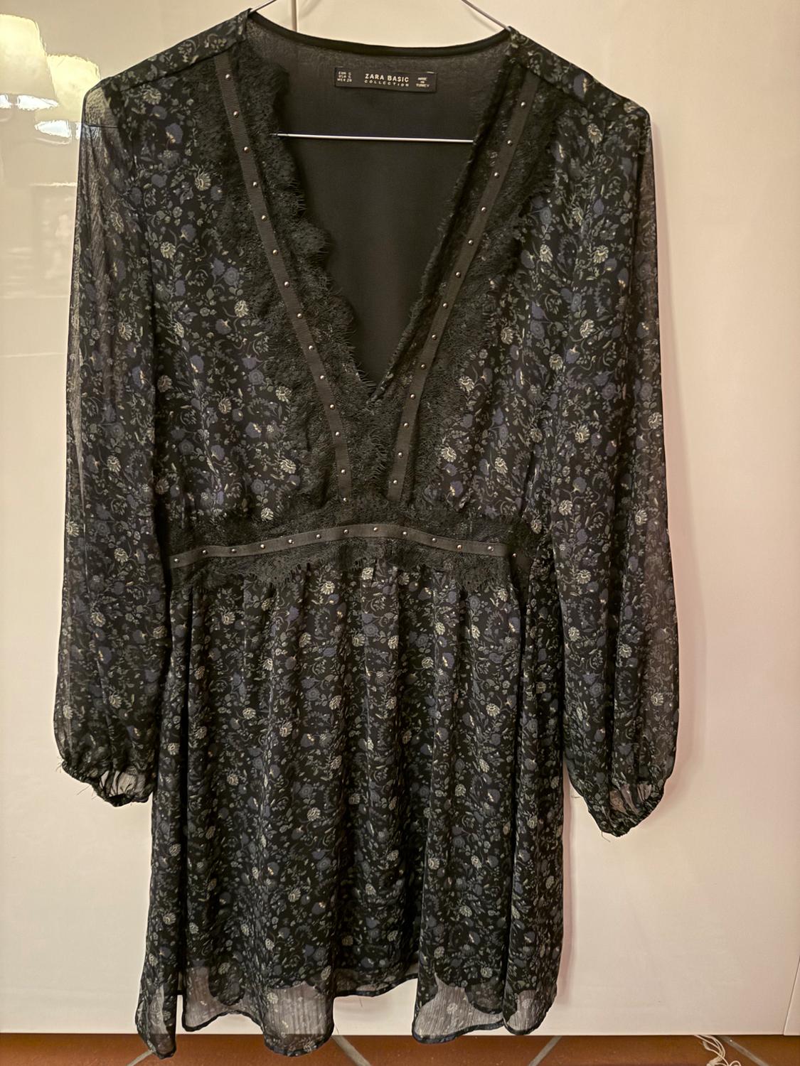Black dress with floral print and lace on the neckline, hanging on a hanger. Long sleeves and small stud details.