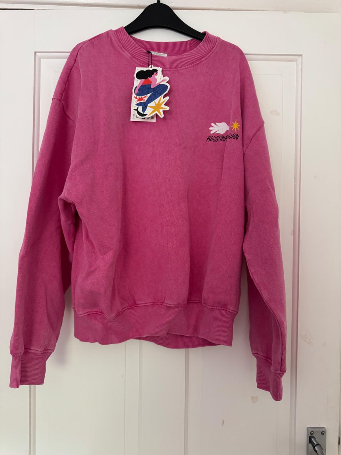 Pink sweatshirt hanging on a white door. It features an illustrated label and a design on the chest with the text AGUSTINA GUERRERO.
