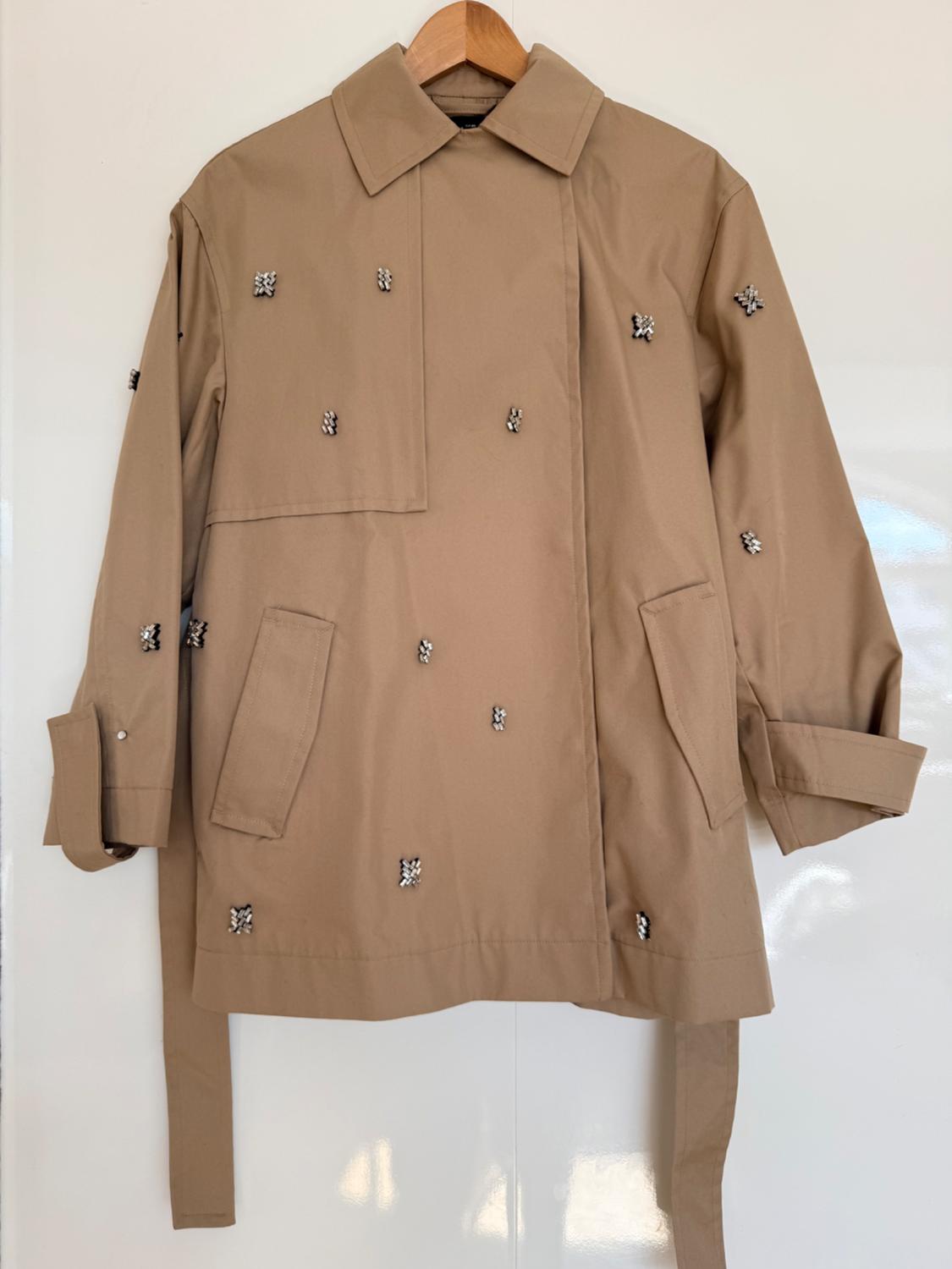 Beige trench coat with rhinestone embellishments, collar and belt, hanging on a wooden hanger against a white background.
