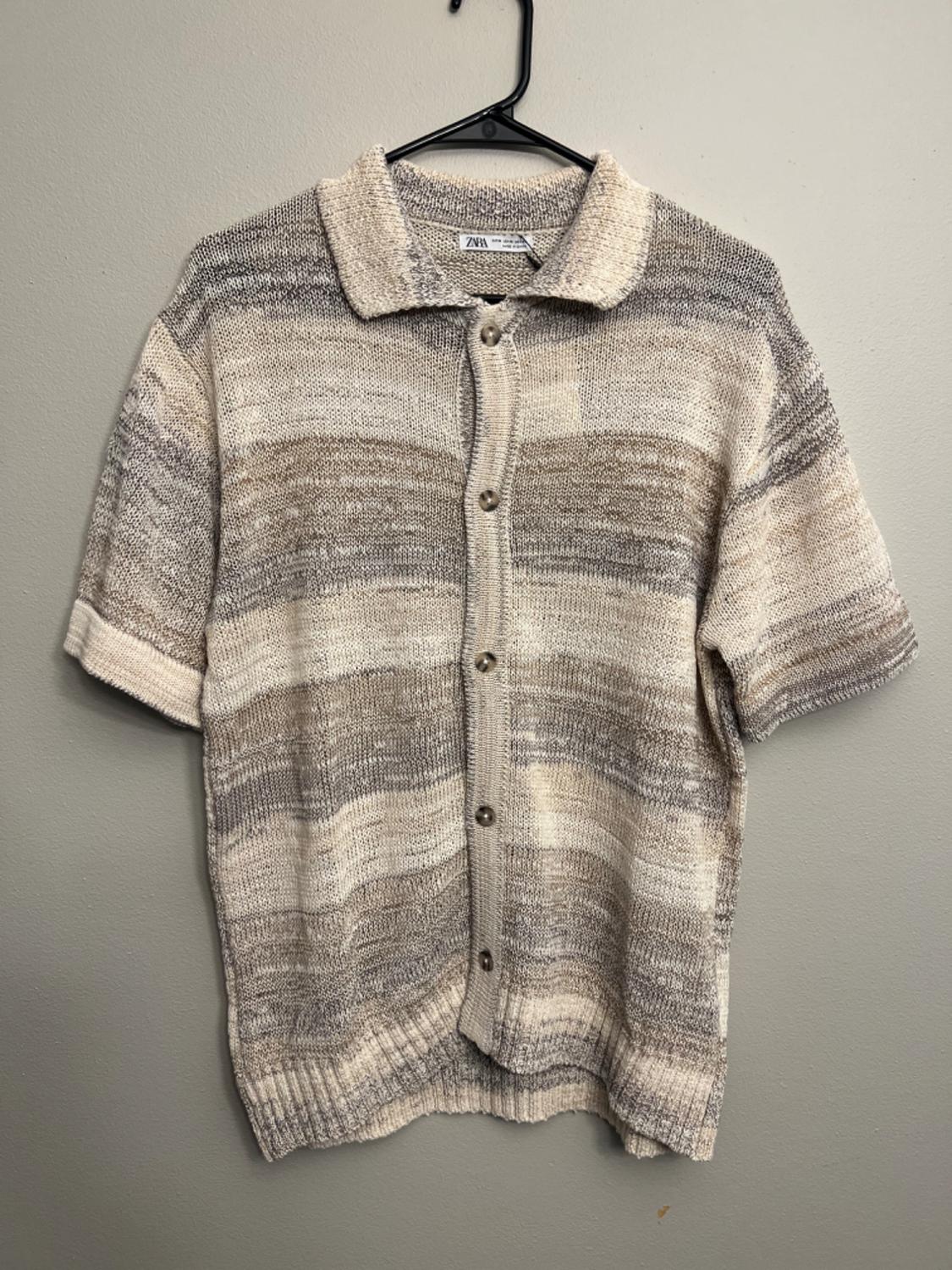 Knit shirt in beige and gray tones with front buttons, hanging on a black hanger on a gray wall. Visible label.