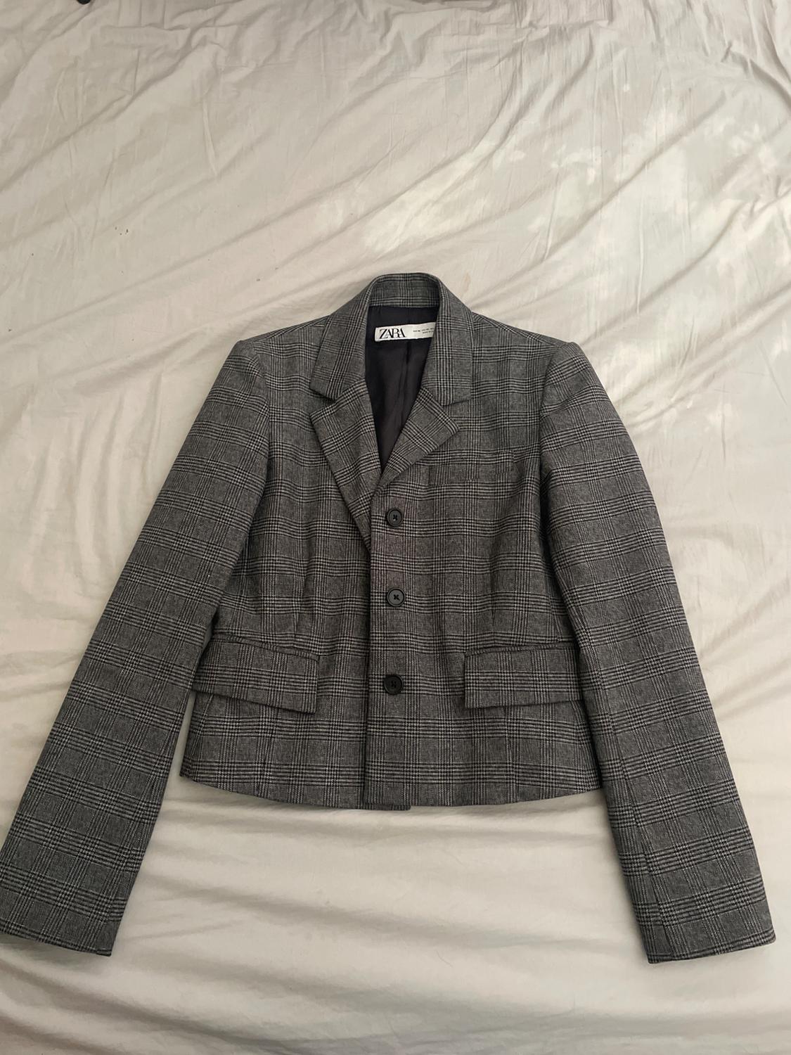 Grey checked jacket with three buttons and lapels, on a light surface. Visible label with the text "ZARA".
