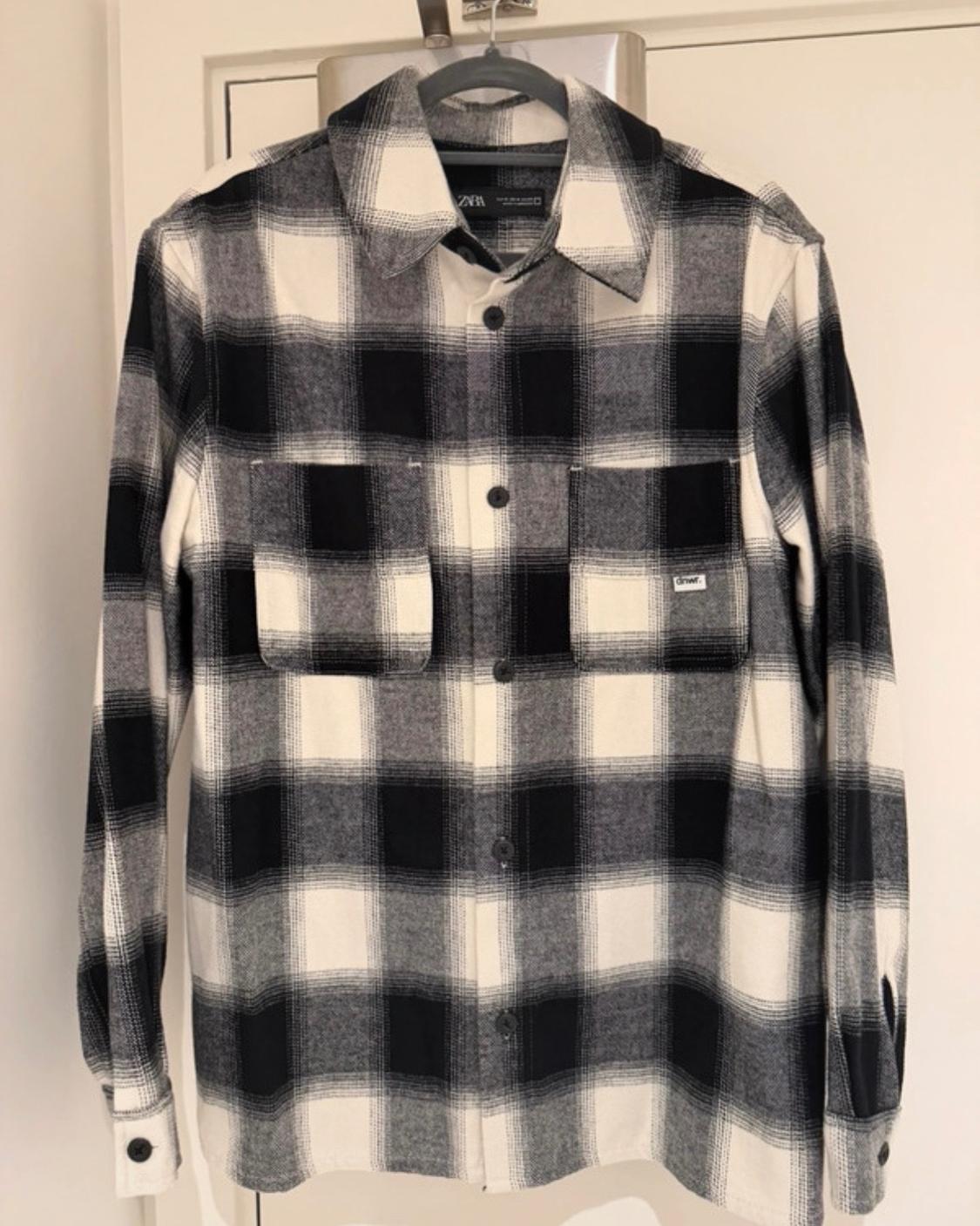 Black, white and grey check shirt hanging on a hanger. It has two front pockets and labels with the text 'ZARA' and 'dnwr.'.
