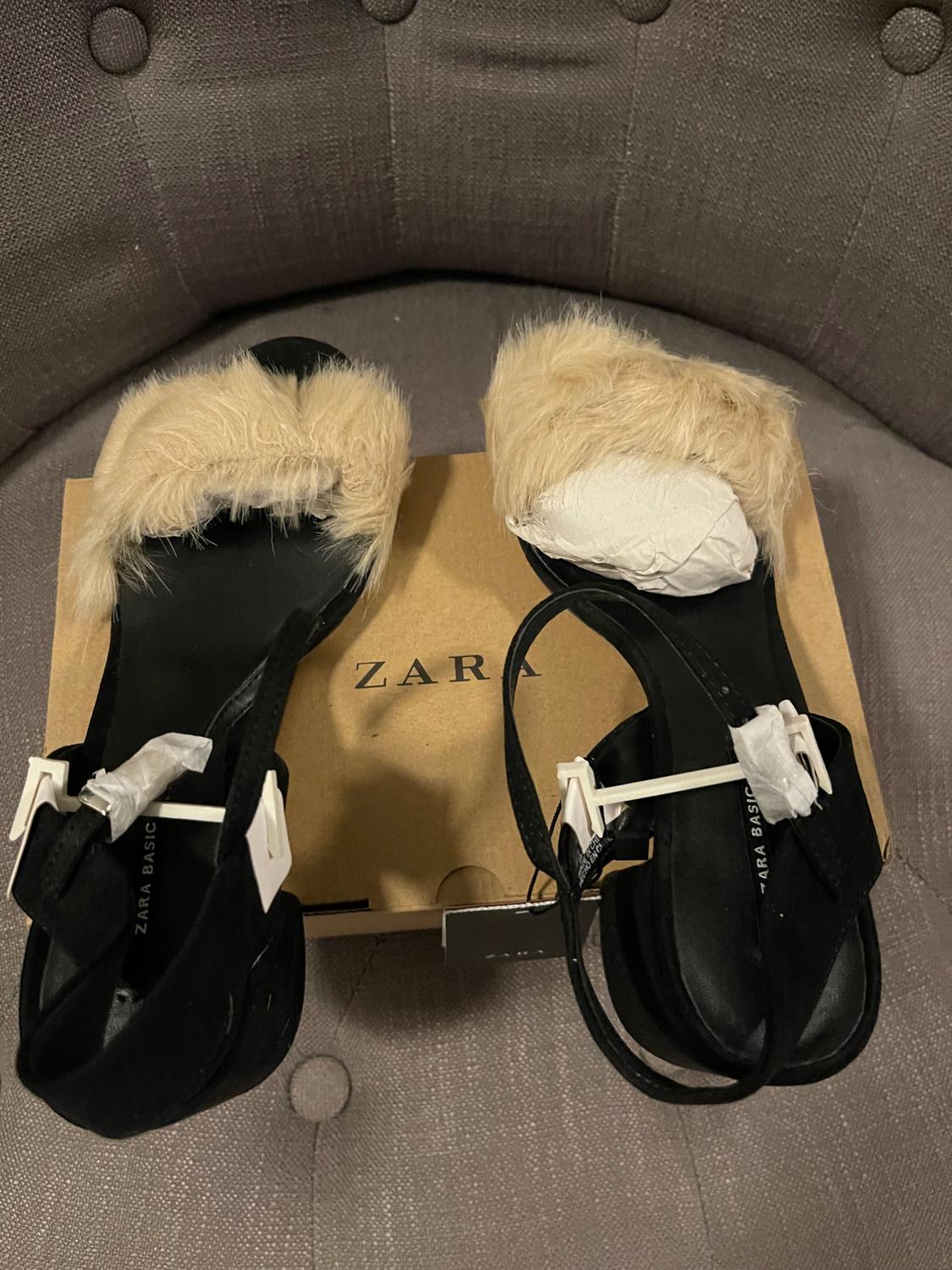 Black sandals with beige fur on a box with the text ZARA. Inside, it reads ZARA BASIC. Composition on a grey armchair.