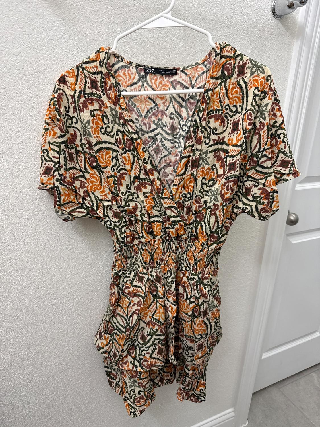 Short dress with a floral print in orange, green, and brown tones, hanging on a white hanger against a white wall.