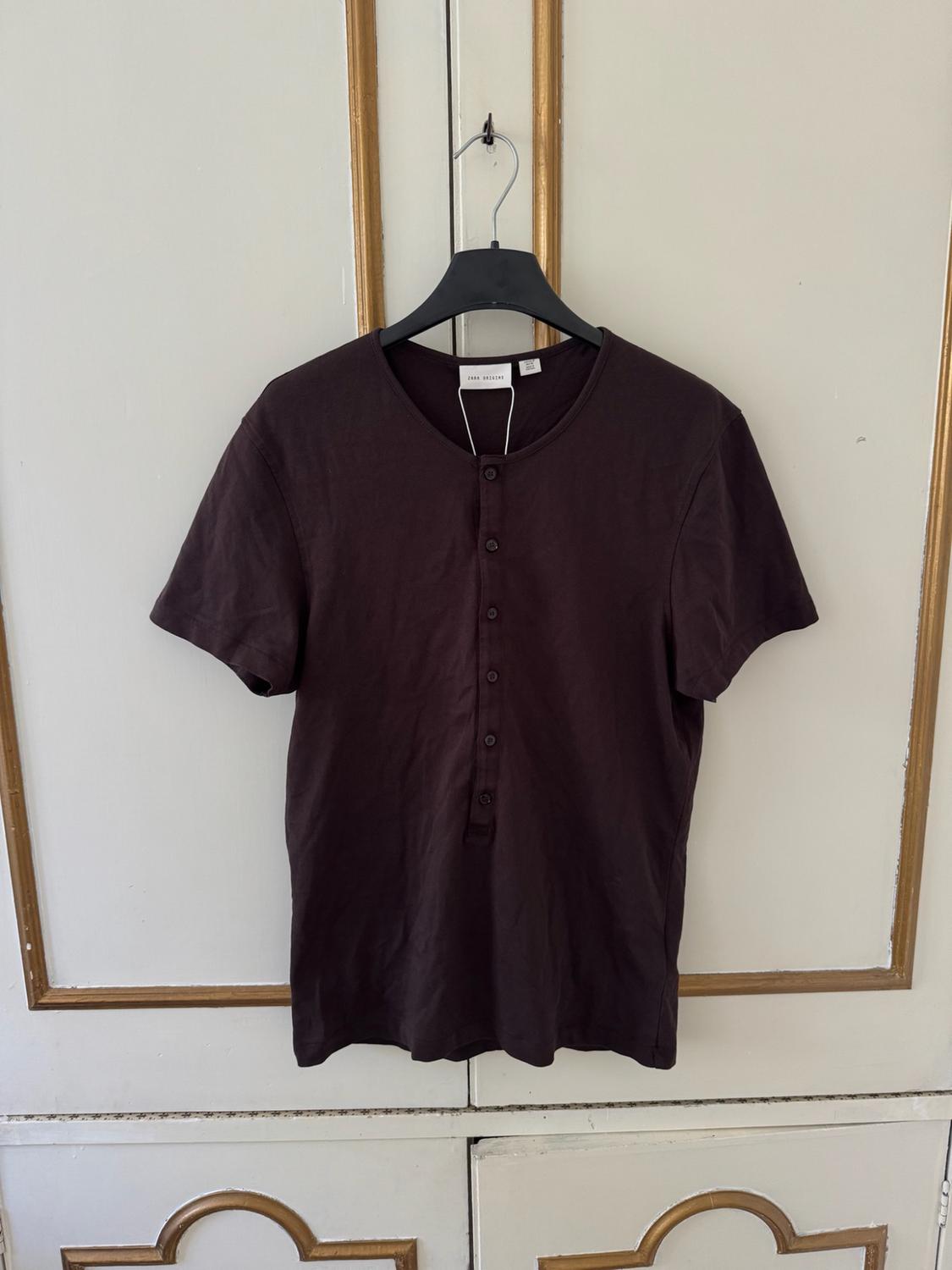 Dark brown short-sleeved T-shirt with buttons at the neck, hanging on a black hanger in front of a white wardrobe with gold details.