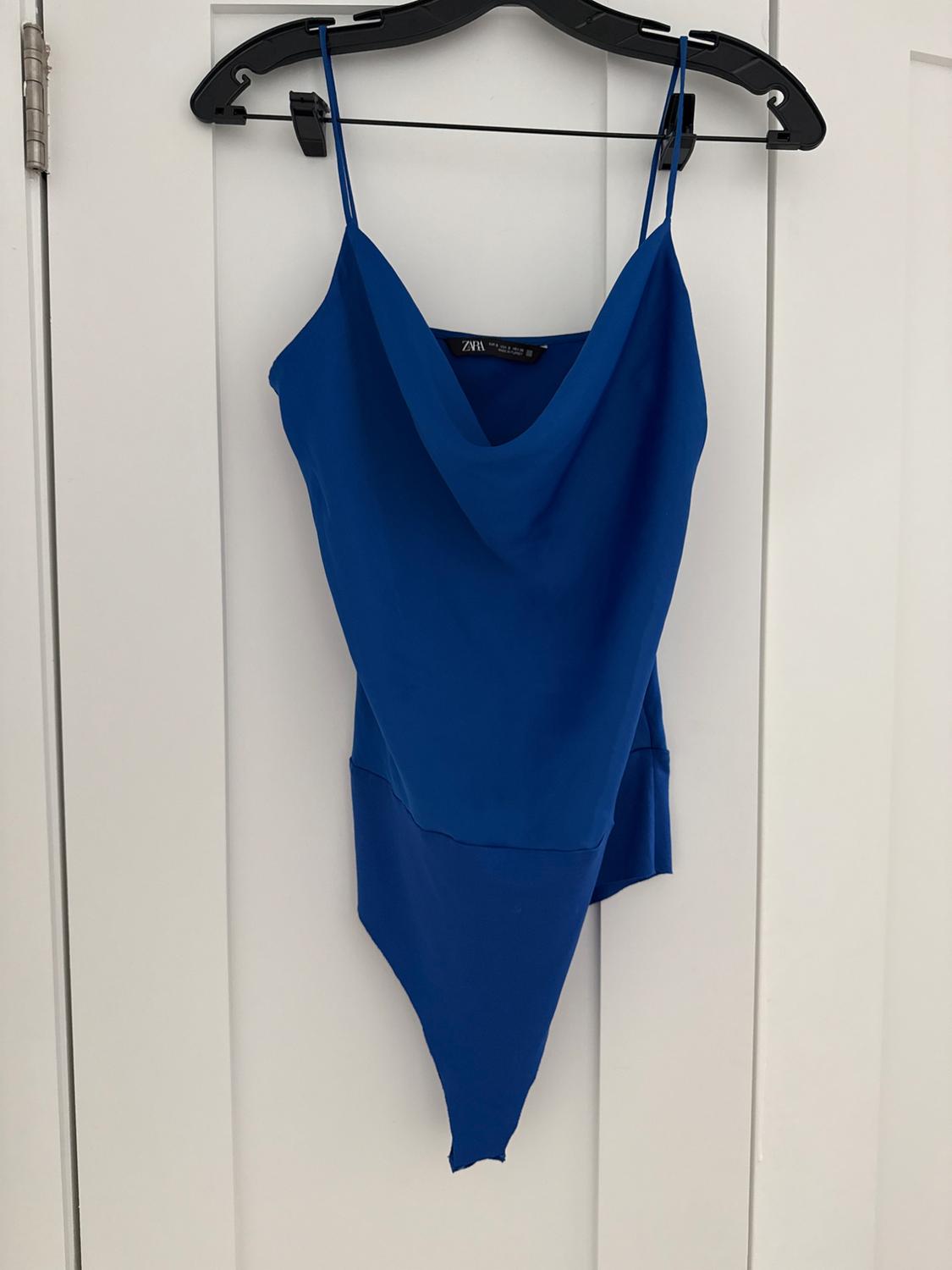 Blue top with thin straps and a draped neckline, hanging on a black hanger over a white door.