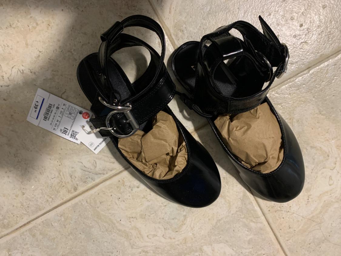 Black patent leather shoes with buckles and paper inside, on a light-coloured floor. Visible tag with a price of 239.