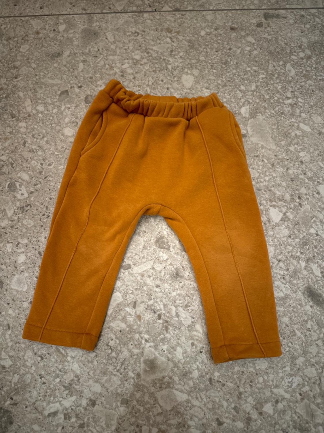 Mustard baby trousers with an elasticated waist, on a textured grey tiled floor.