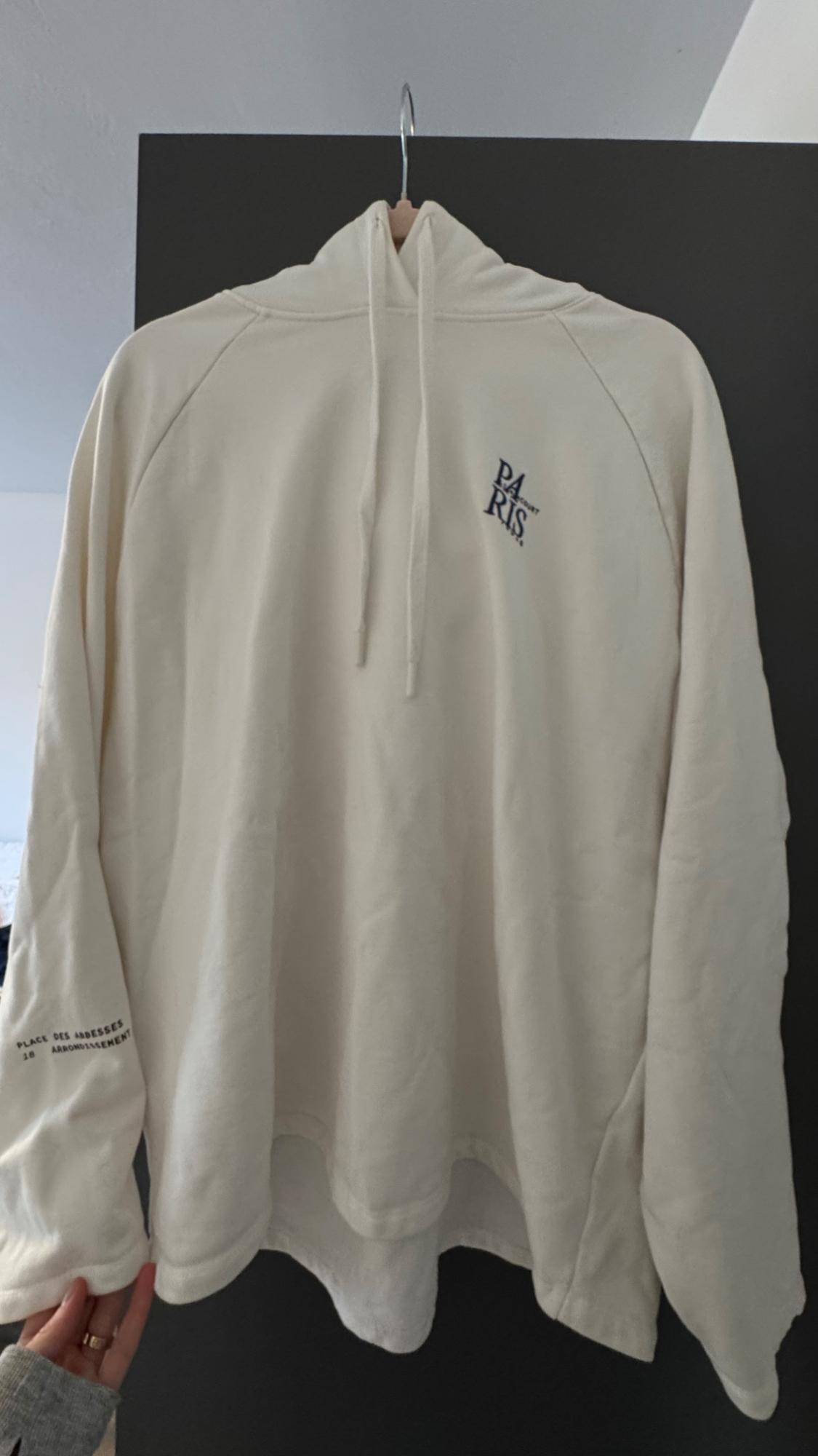 White hooded sweatshirt hanging on a hanger. Visible text: "PARIS" on the chest and "PLACE DES ABBESSES 18 ARRONDISSEMENT" on the sleeve.
