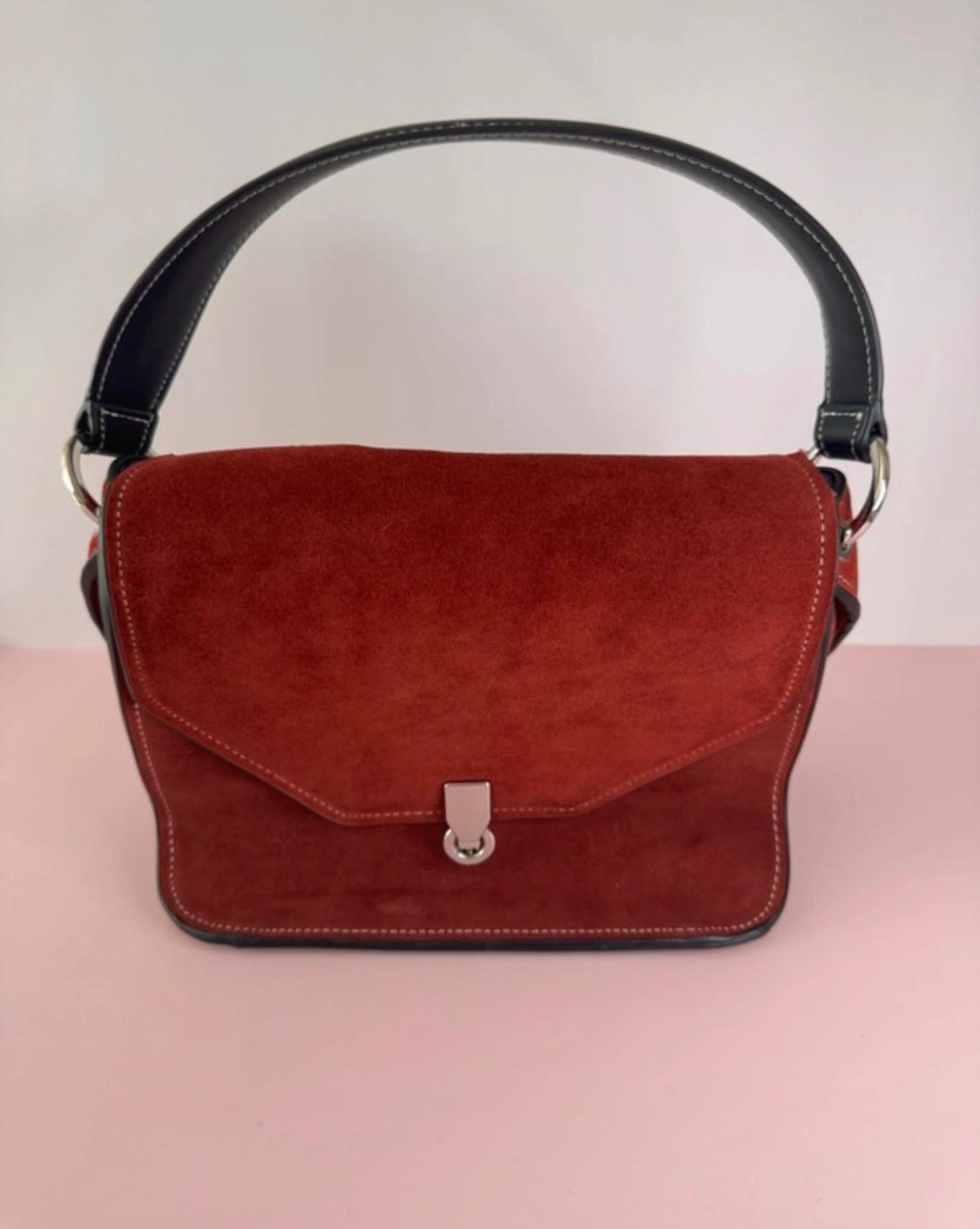 Brown handbag with flap and metallic clasp, black handle. Light pink background.
