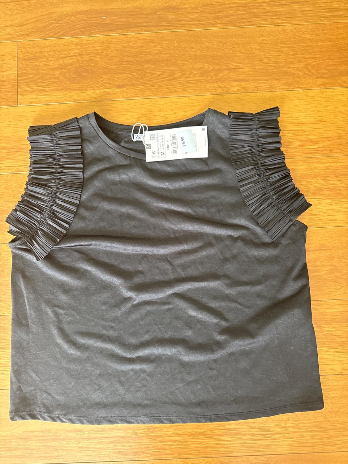 Black T-shirt with pleated sleeves on a wooden floor. Visible label with barcode and price of 32.90.