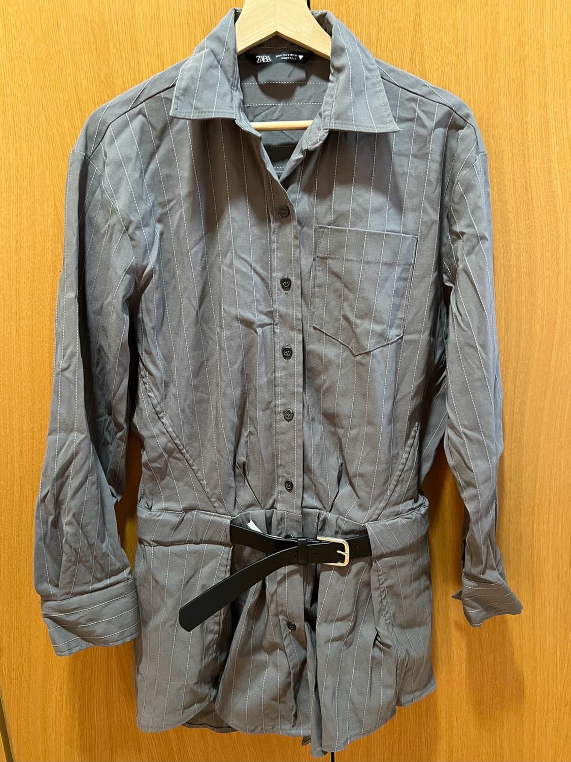 Grey long-sleeved shirt dress with white stripes, black belt and ZARA label, hanging on a hanger against a wooden background.