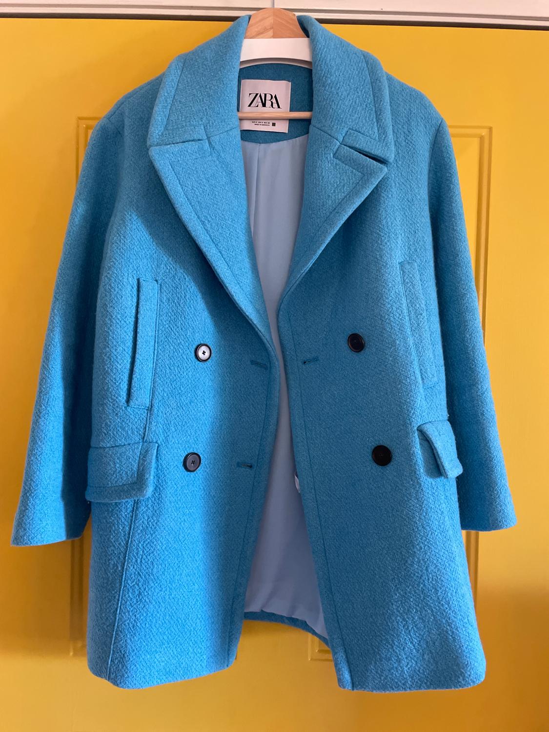 Blue coat hanging on a yellow door. The coat has black buttons and a label that says "ZARA".
