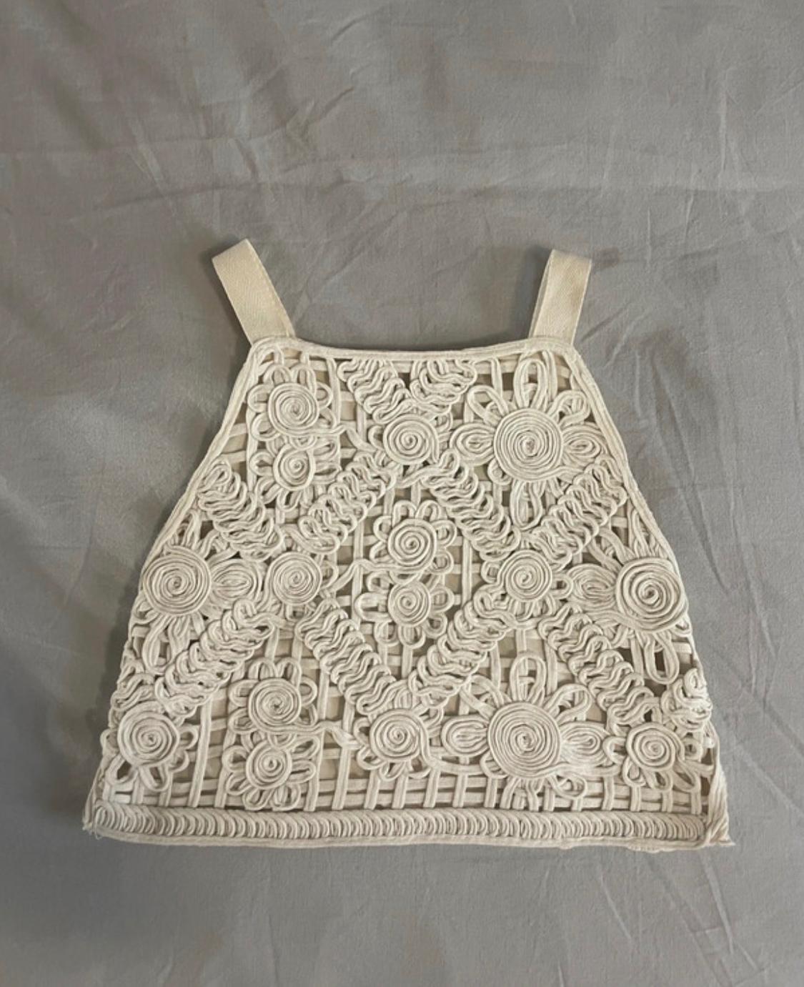 Ecru macramé top with floral motifs and wide straps, against a grey fabric background.