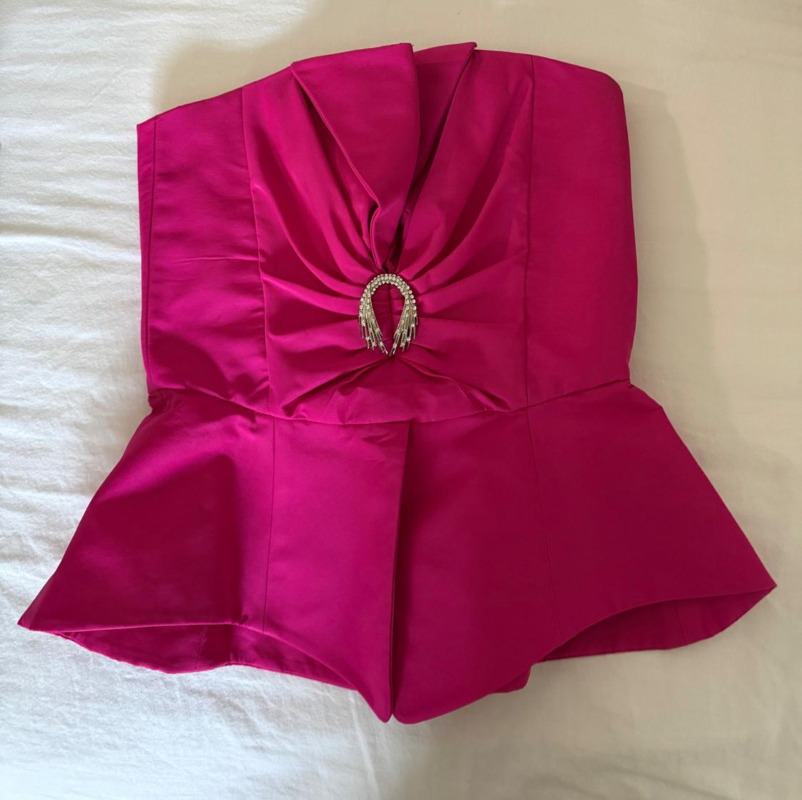 Fuchsia strapless top with front pleat detail and horseshoe-shaped metallic embellishment, on a white background.