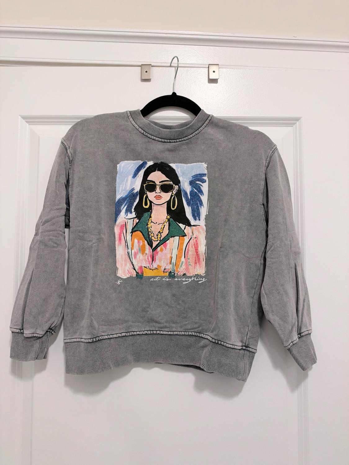 Gray sweatshirt hanging on a white door. It features a print of a woman with sunglasses and the text "art is everything".