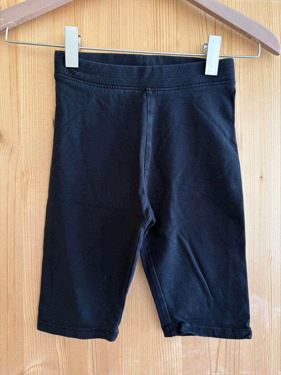 Black short leggings hanging from a wooden hanger with metal clips against a light wood background. Relaxed, casual style.